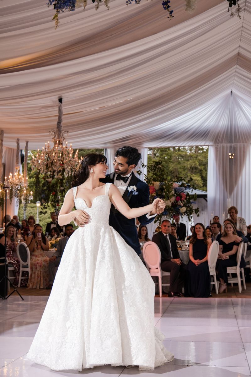 A bride and groom share their first dance at their wedding reception, surrounded by seated guests under a white draped ceiling with chandeliers.