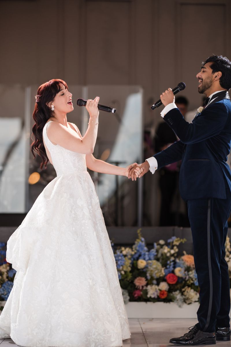 A bride and groom hold hands and sing into microphones during their wedding reception