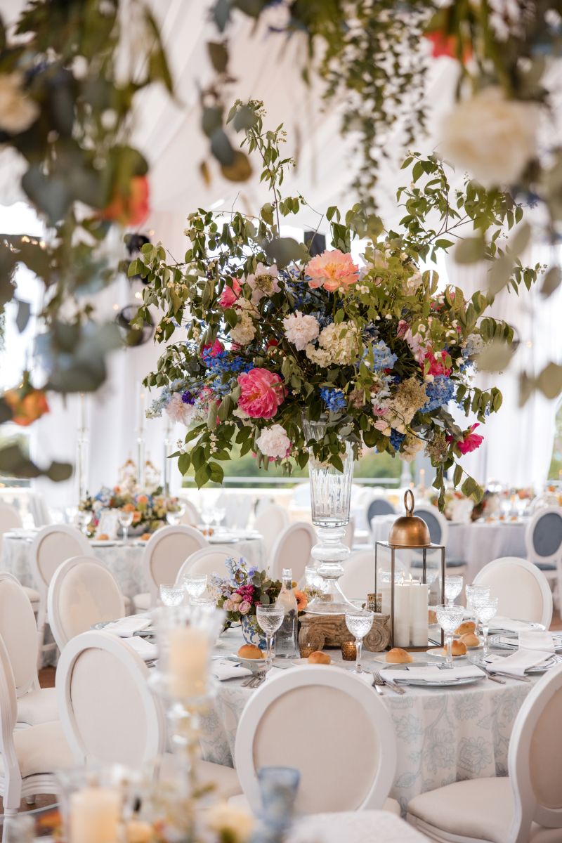 Elegant banquet tables are set with white chairs, glassware, and lanterns, featuring a tall floral centerpiece with colorful flowers and greenery