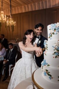 A bride and groom, dressed formally, cut a white tiered wedding cake adorned with blue and white flowers