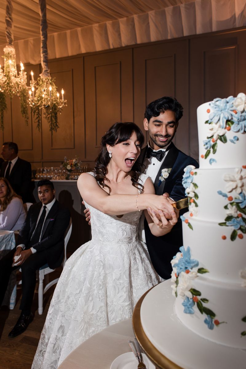 A bride and groom, dressed formally, cut a white tiered wedding cake adorned with blue and white flowers