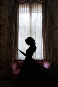 Silhouette of a woman in a dress standing before a large window with sheer curtains and ornate drapes, next to a pink upholstered sofa