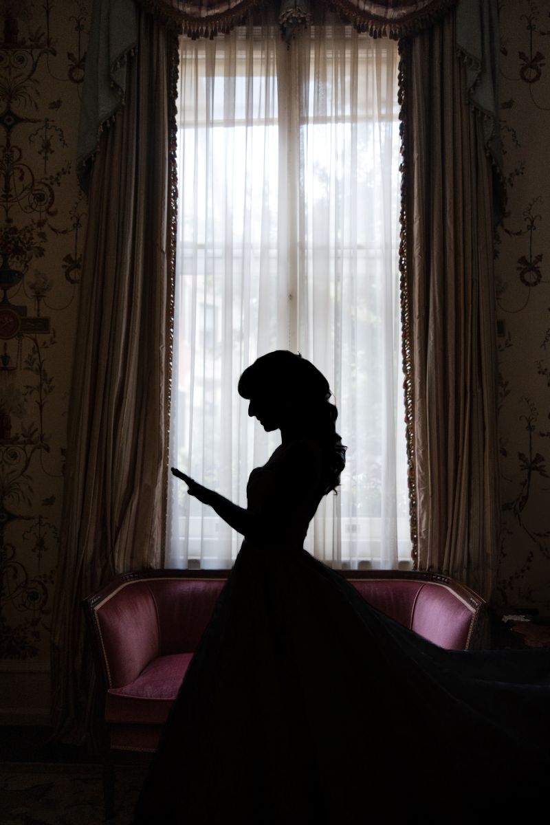 Silhouette of a woman in a dress standing before a large window with sheer curtains and ornate drapes, next to a pink upholstered sofa