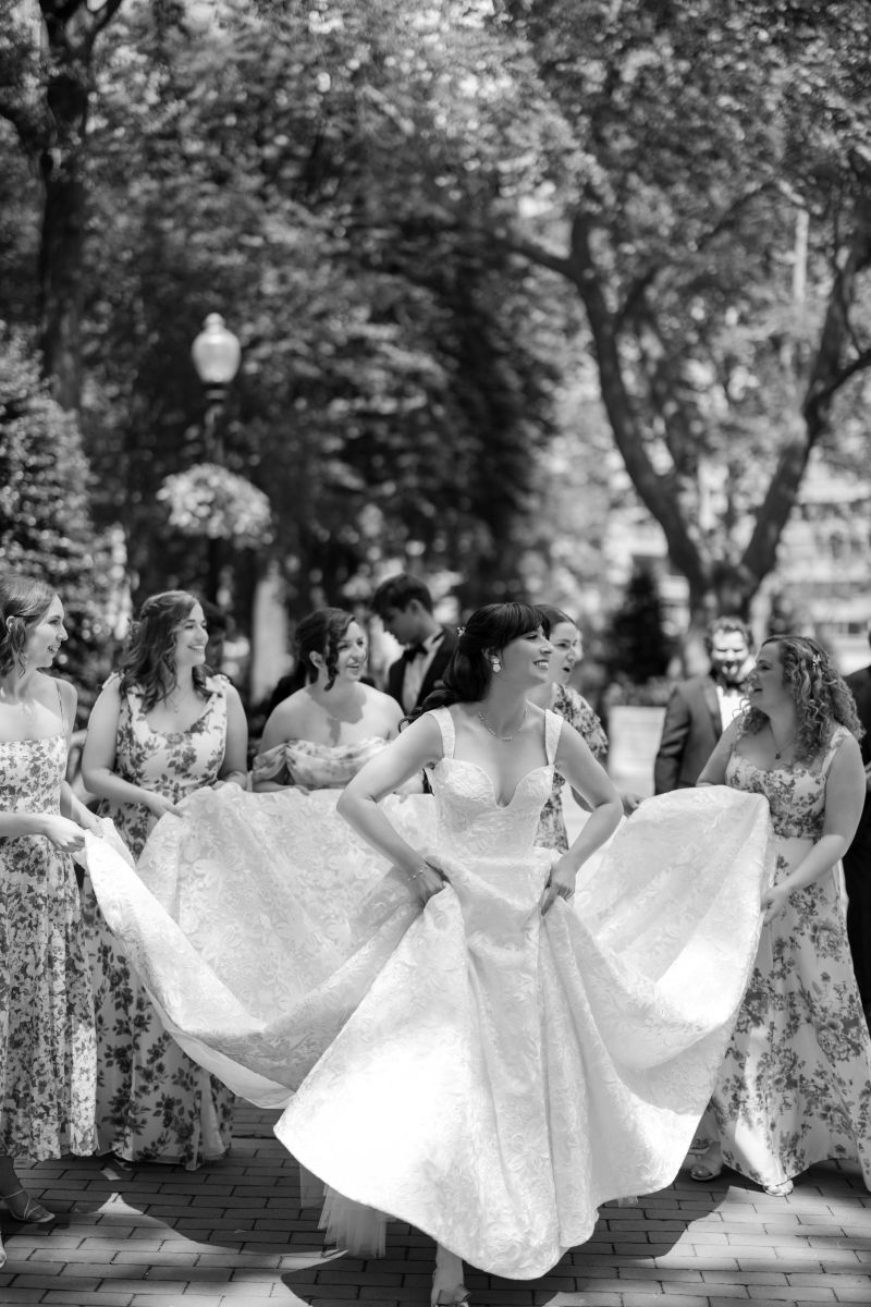 A bride in a white dress stands smiling with her bridal party, all wearing floral dresses, outdoors on a sunny day