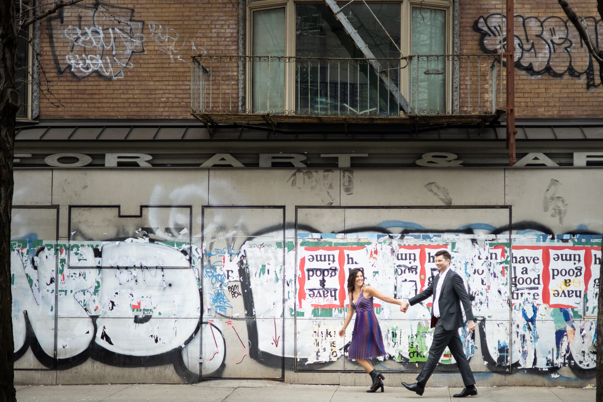 A woman in a dress and a man in a suit walk hand-in-hand past a graffiti-covered wall and posters on a city sidewalk, capturing candid New York City engagement photos.