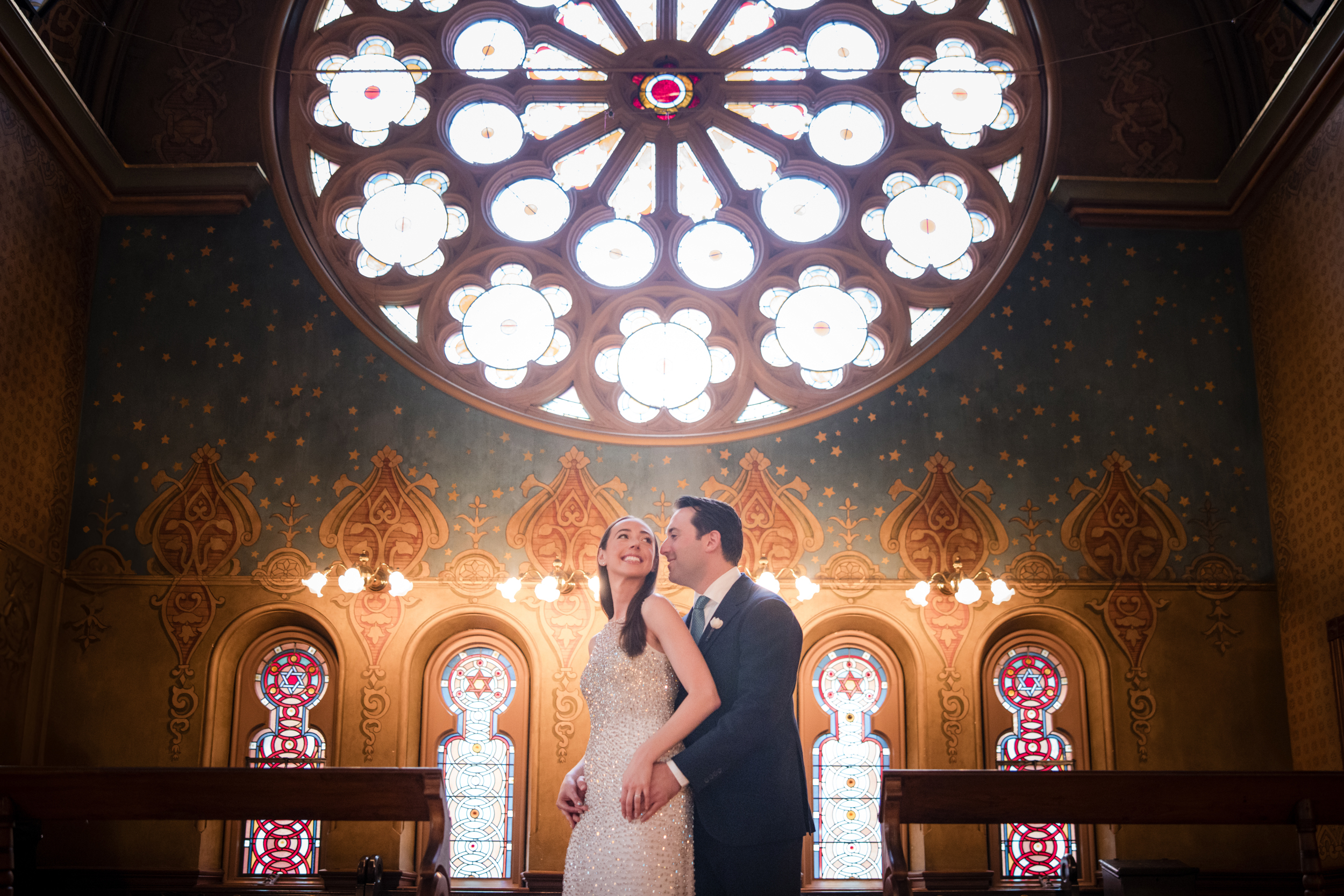 A bride and groom stand together, smiling, in front of decorative stained glass windows inside the Museum at Eldridge Street.