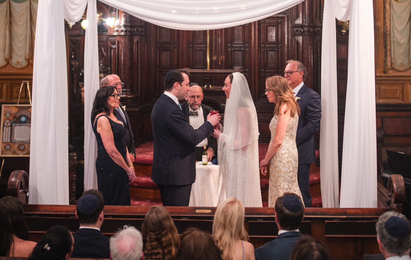 A wedding ceremony takes place under a white chuppah in the ornately decorated Museum at Eldridge Street, with guests seated and musicians playing nearby.