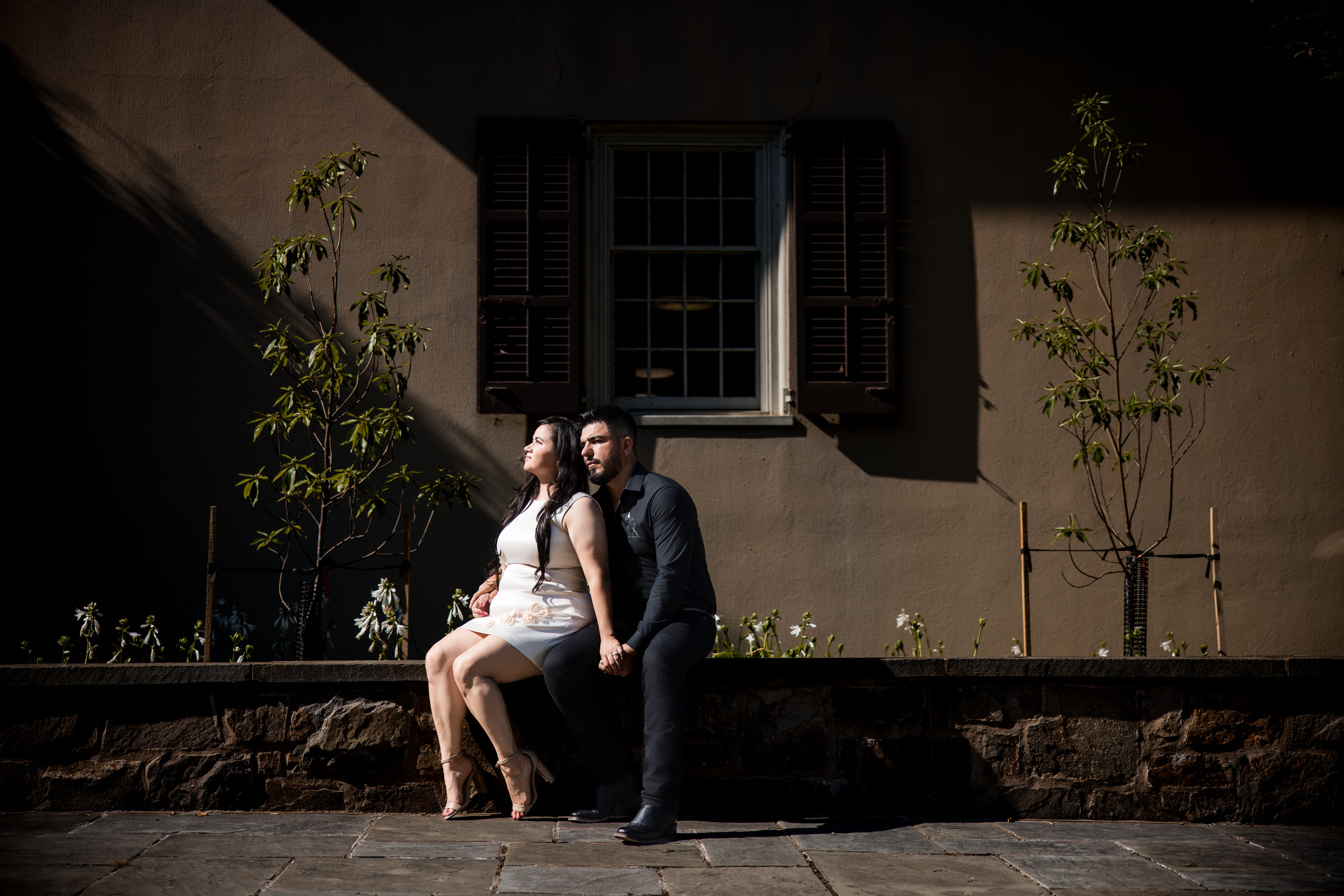 A woman in a white dress and a man in dark clothing sit back-to-back on a stone ledge outdoors, with a building, window, and small trees in the background at a Winterthur engagement photo shoot
