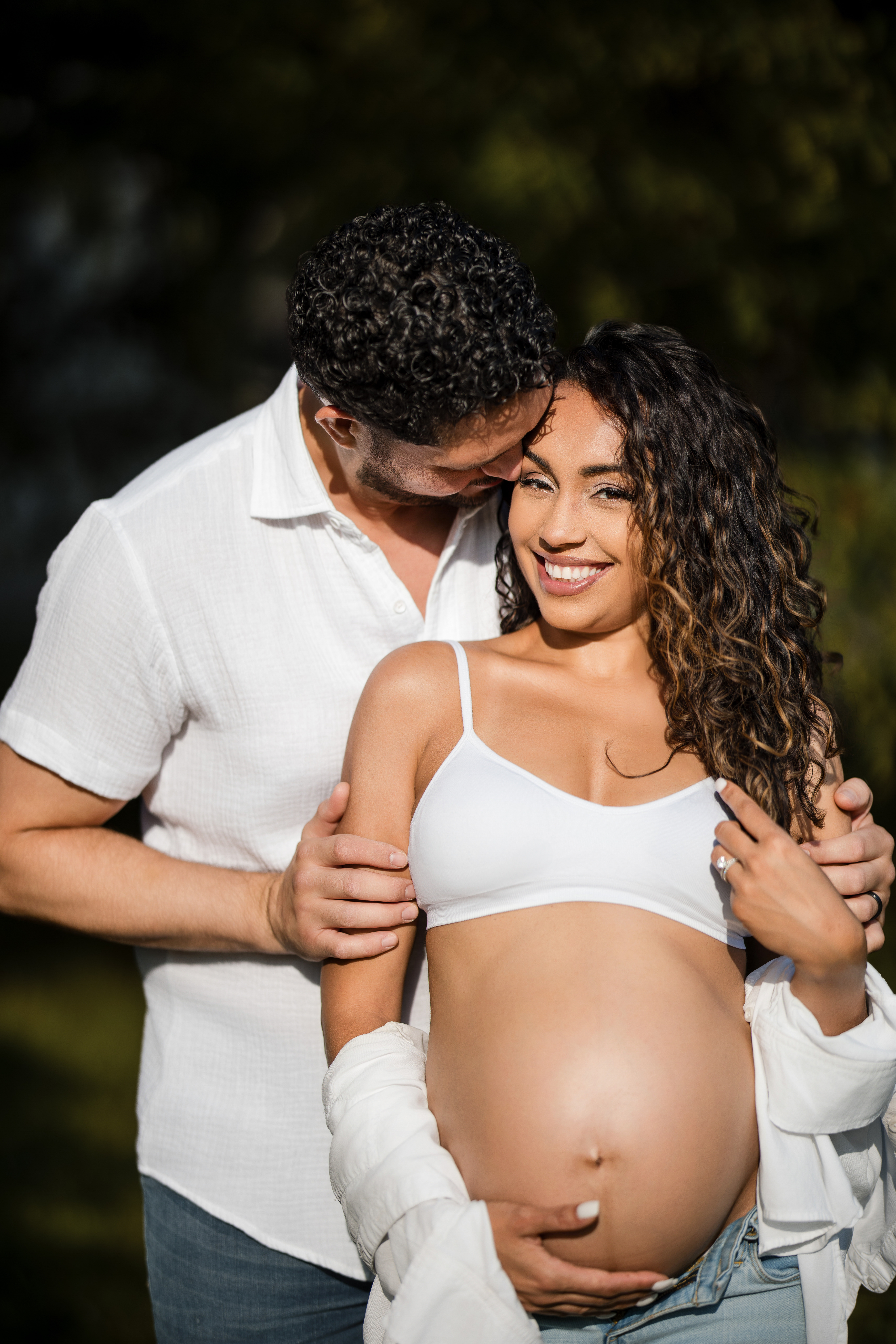 A man stands behind a smiling pregnant woman with his hands on her belly during a maternity session at Valley Forge Park. They are outdoors, and the woman is wearing a white top and open shirt.