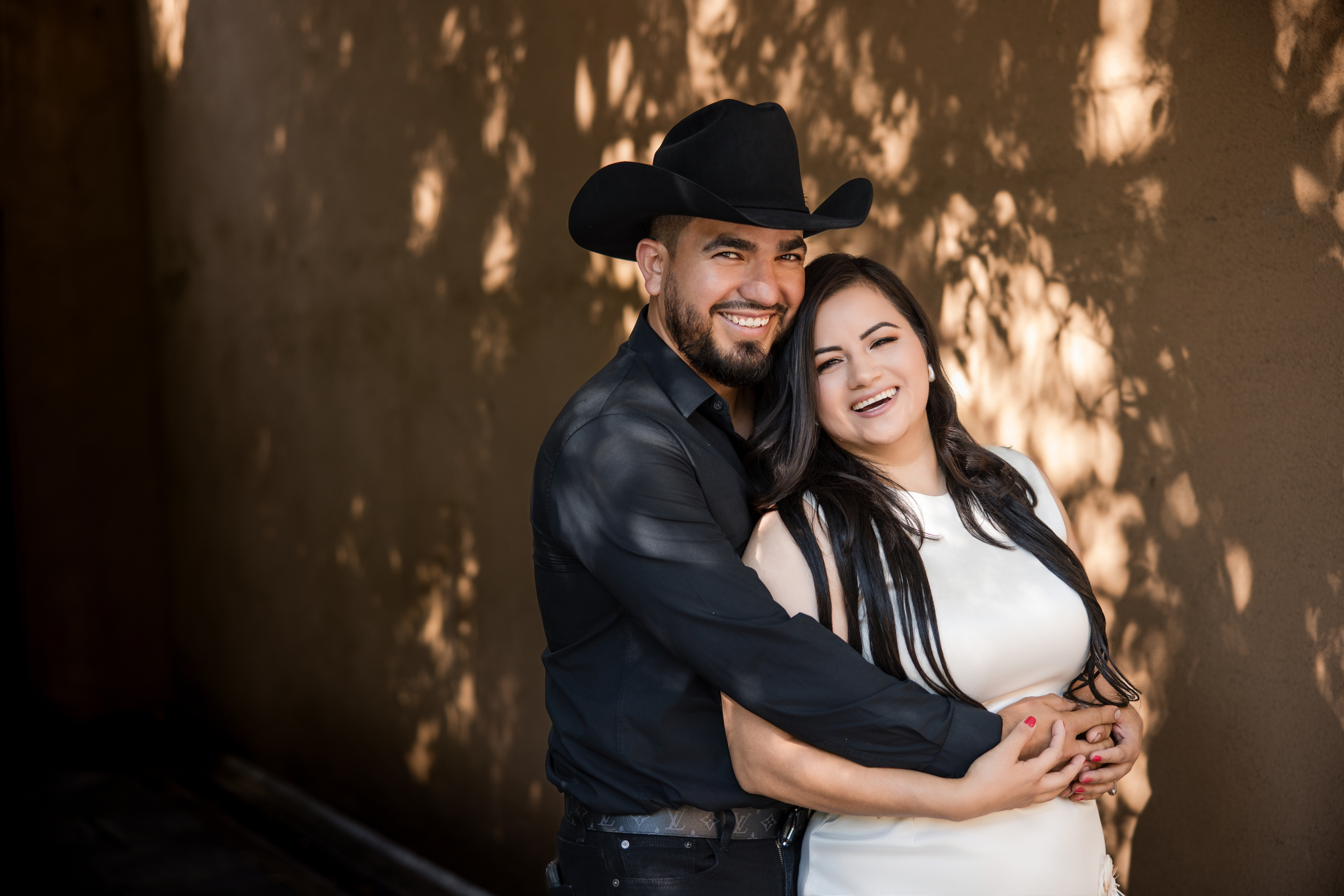 A man in a black cowboy hat and a woman in a white dress stand close together, smiling and embracing in front of a sunlit wall with leafy shadows, capturing the warmth of their Winterthur engagement photos.