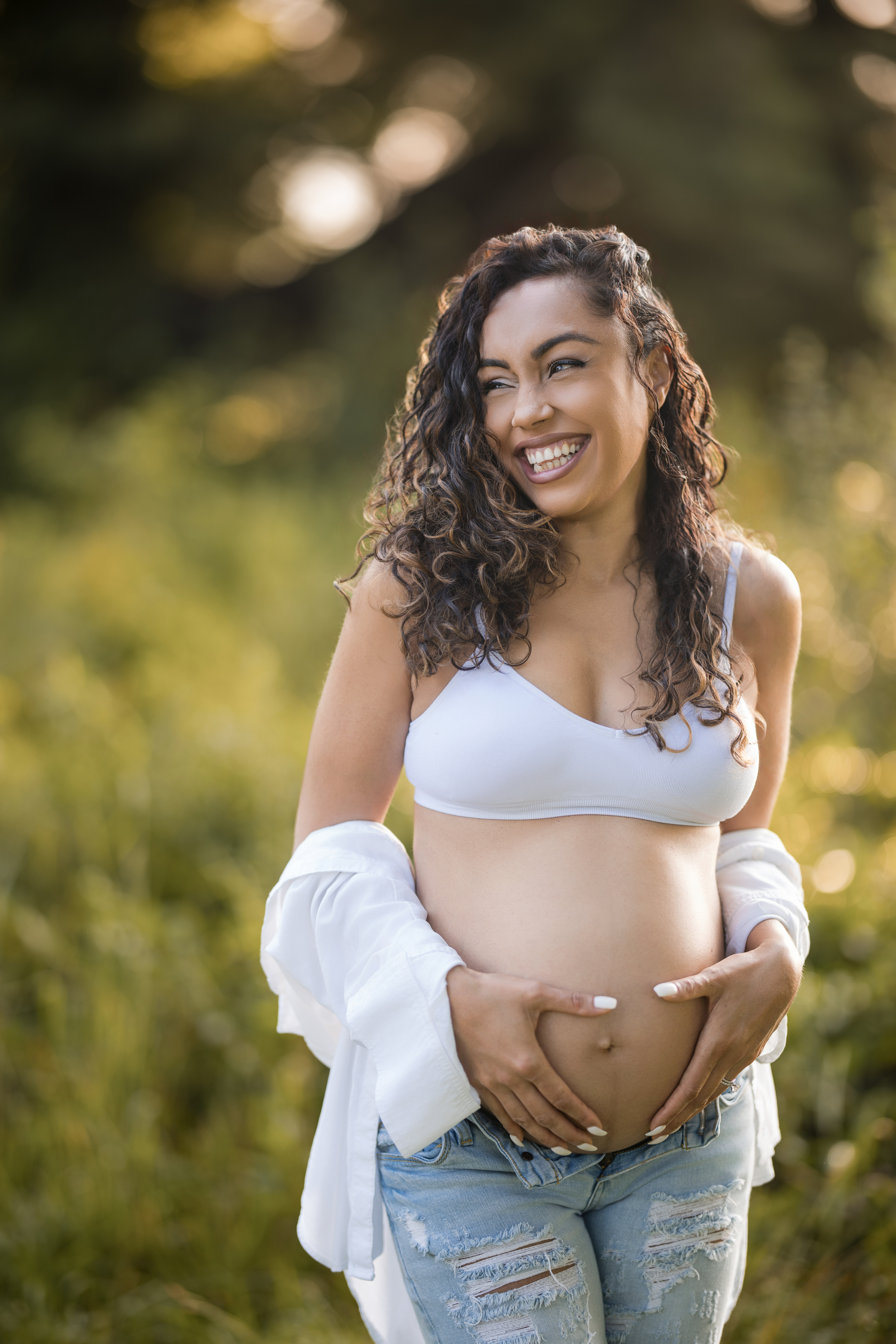A pregnant woman in a white bra and ripped jeans stands outdoors, smiling and holding her belly, during a maternity session at Valley Forge Park, with lush greenery in the background.