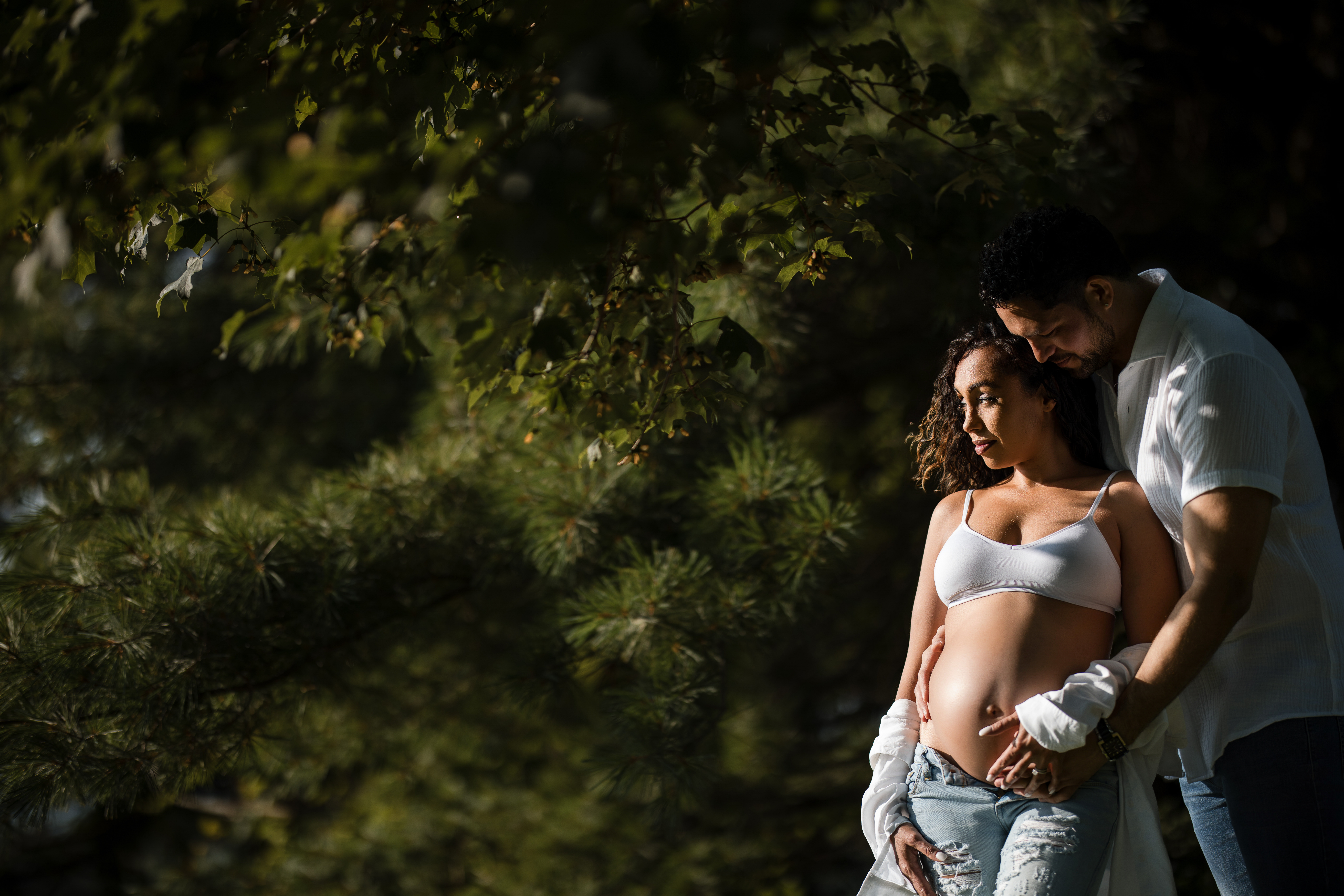 A couple stands outdoors during their maternity session at Valley Forge Park, with the man gently holding the woman’s pregnant belly. Both are dressed casually, surrounded by greenery and sunlight filtering through trees.