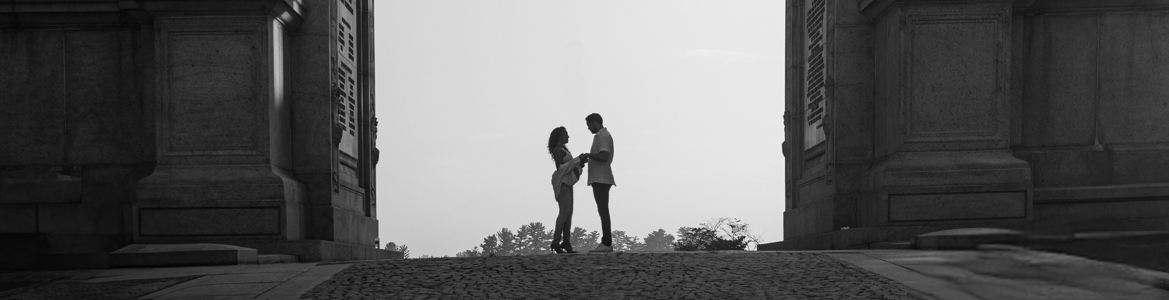 Two people stand beneath a large stone arch monument, silhouetted against the sky, with trees on the right—a touching moment from a maternity session at Valley Forge Park.