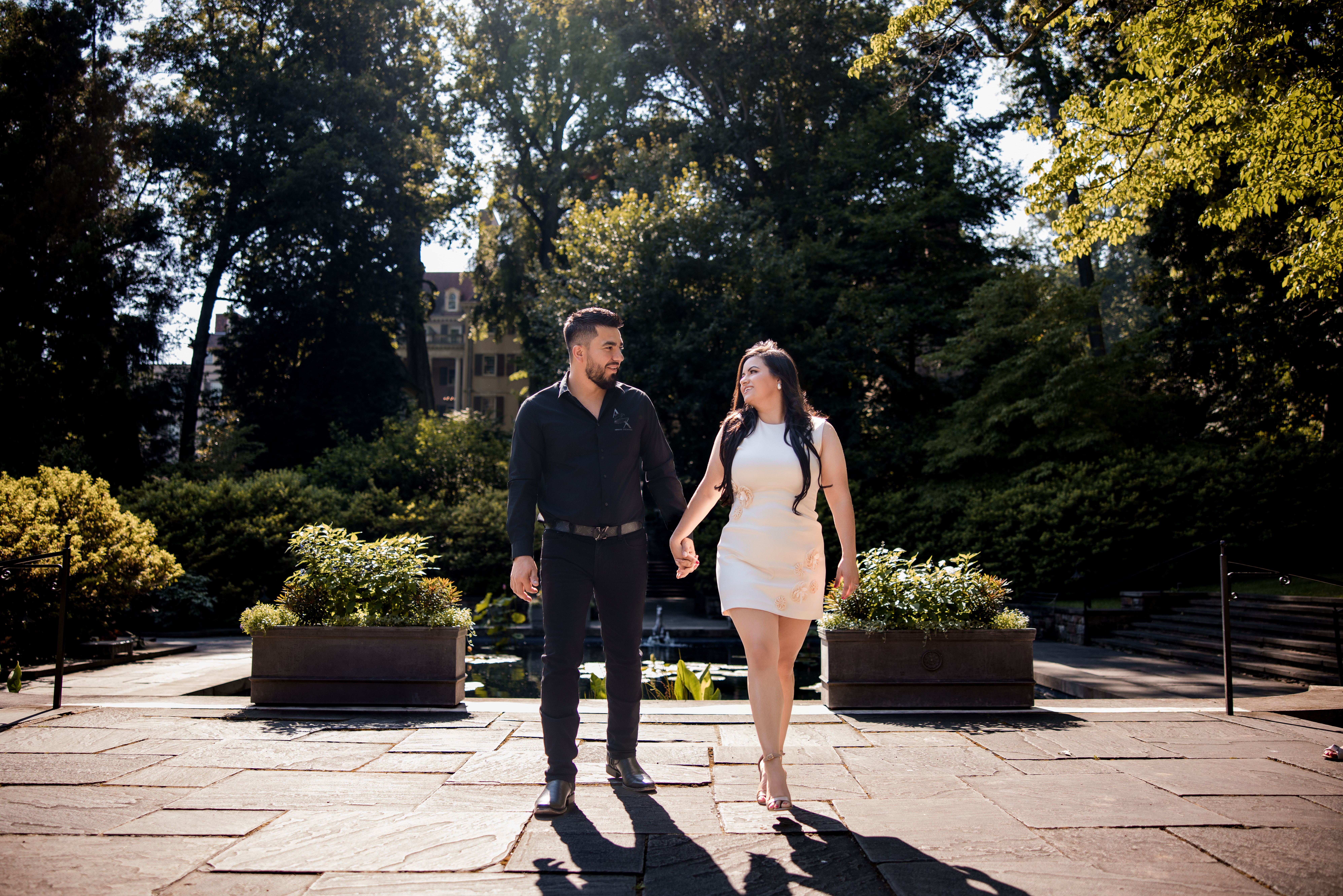 Engaged couple walking hand-in-hand through the Reflecting Pool Garden at Winterthur Museum in Delaware during their summer engagement session, surrounded by formal stone paths and lush greenery.