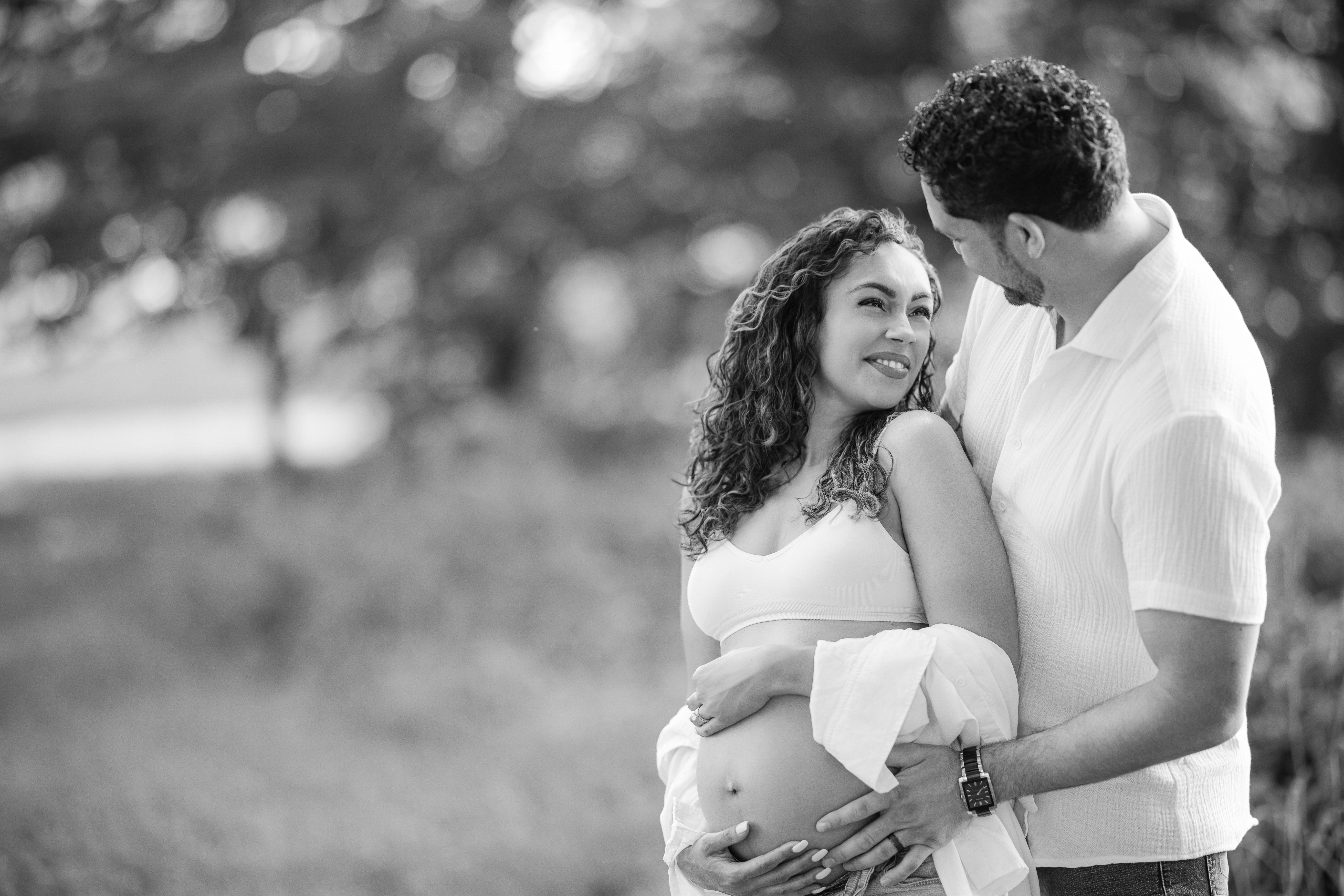 A couple stands outdoors during a maternity session at Valley Forge Park, with the man embracing the smiling pregnant woman from behind as they both cradle her bare belly.