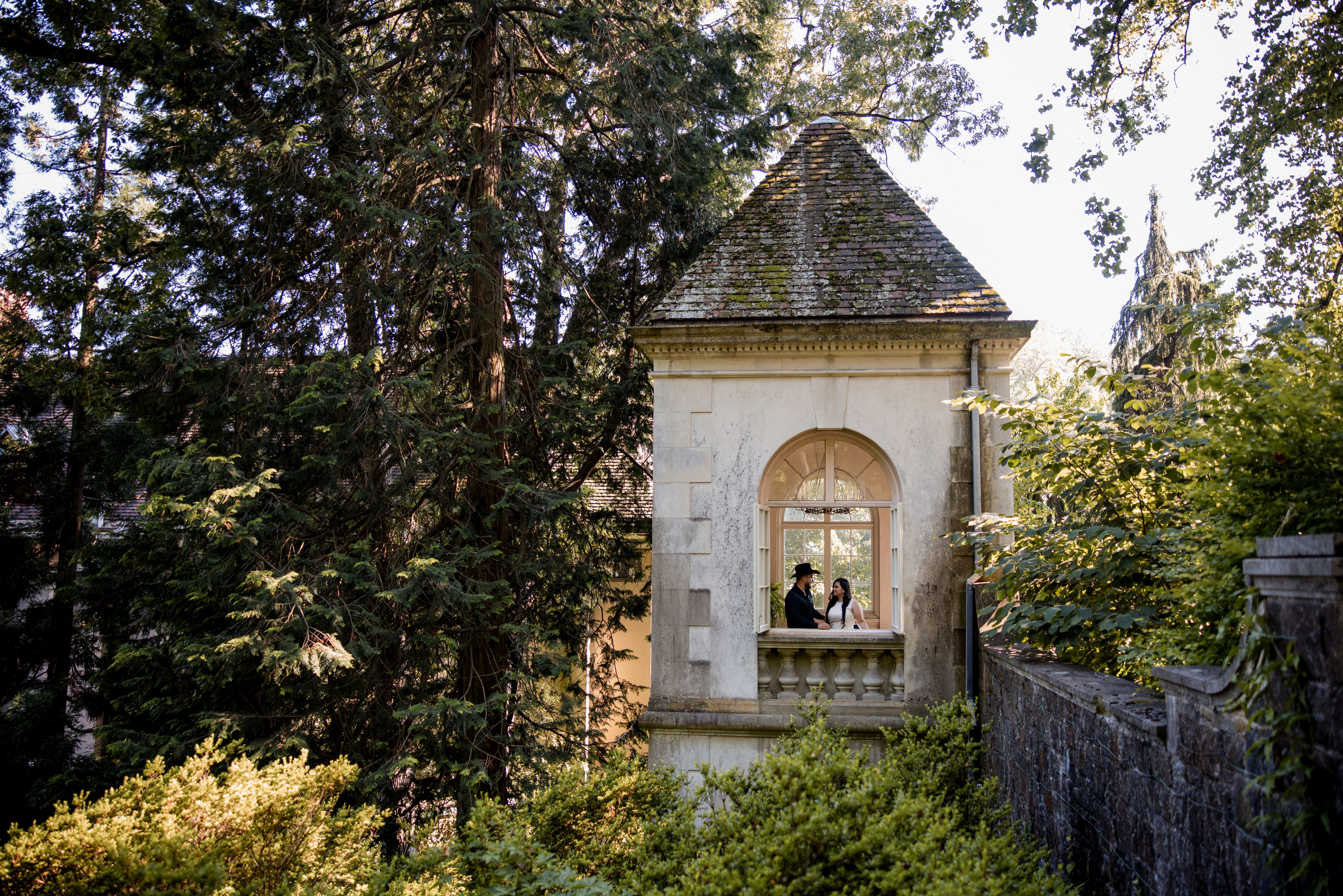 Couple standing on the stone balcony of a historic pavilion at Winterthur Museum in Delaware during their engagement session, surrounded by tall trees and lush greenery. 