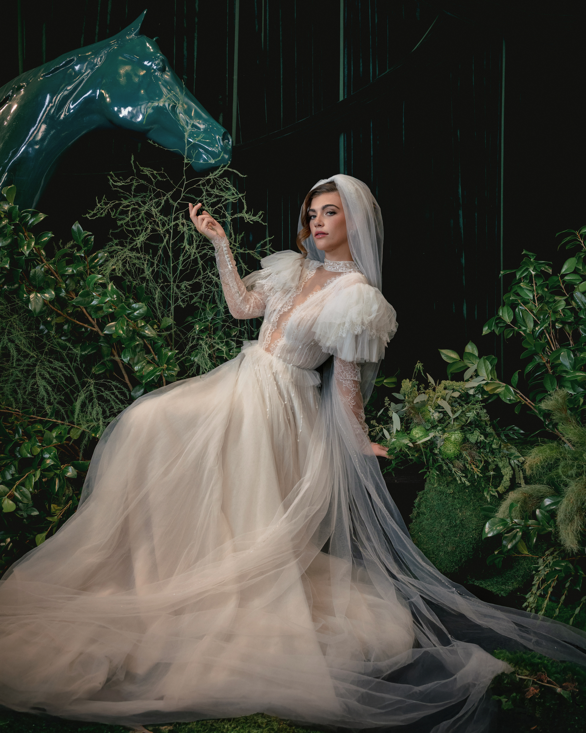 A woman in a white wedding dress and veil sits surrounded by greenery, with a blue sculpted horse head in the background—capturing the artistic essence of Editorial Wedding Photography in NYC.