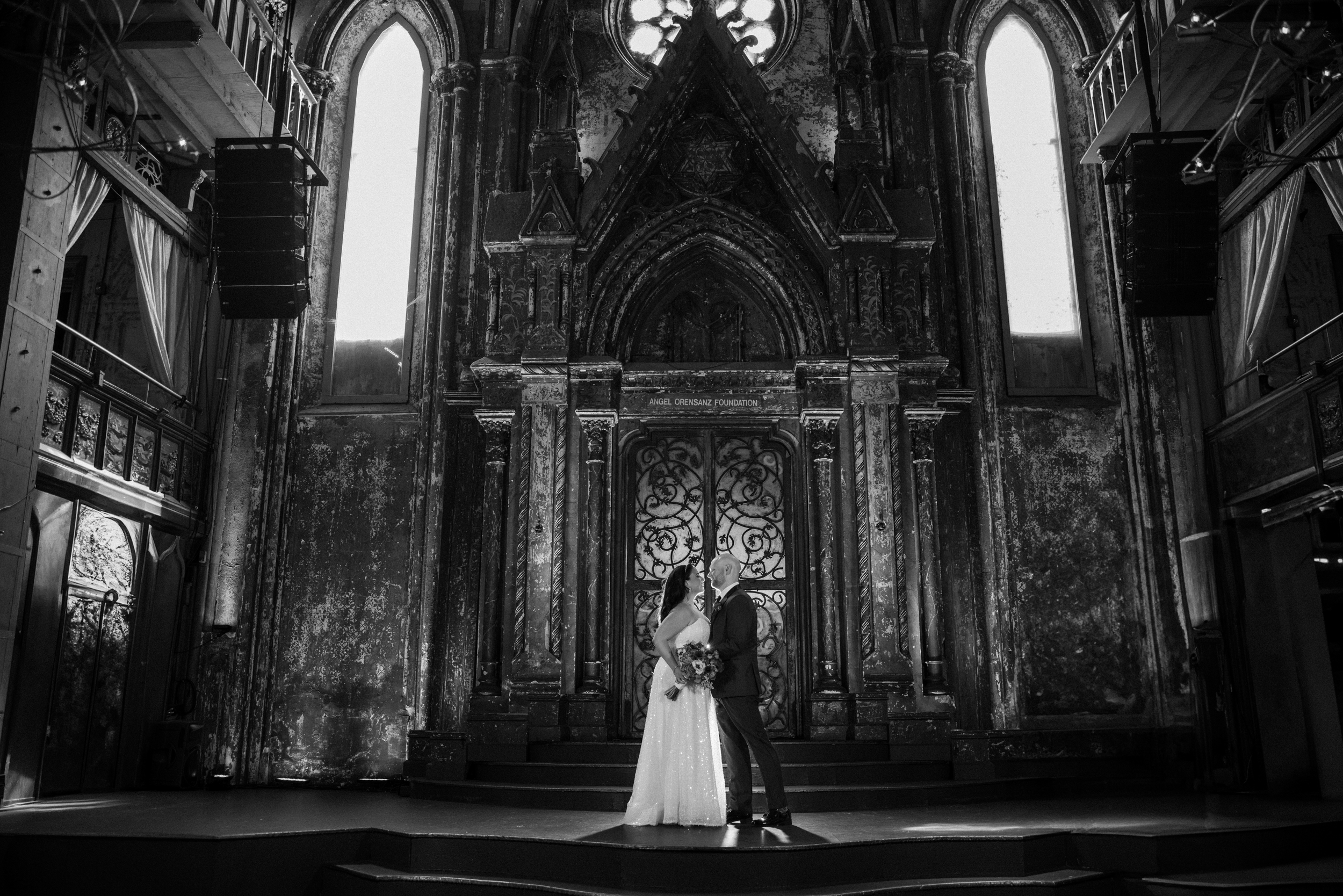 A bride and groom stand together onstage in an ornate, gothic-style venue with tall arched windows and decorative metal doors behind them, capturing the enchanting atmosphere of an Angel Orensanz Wedding.