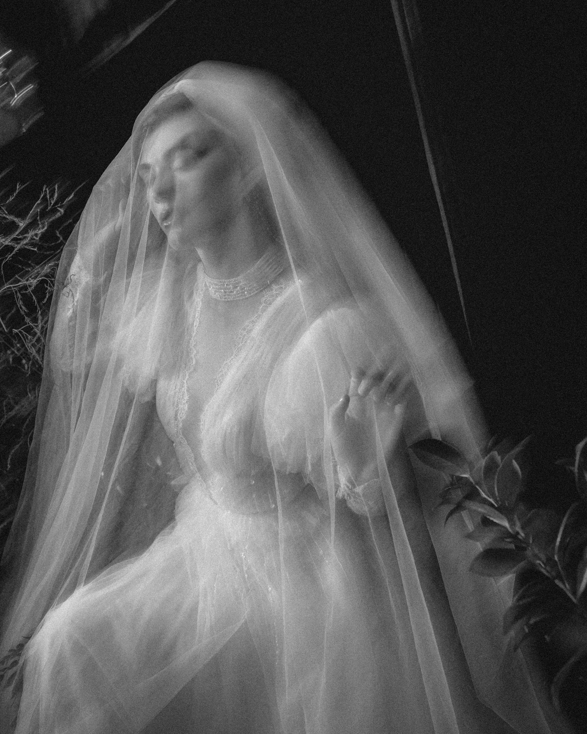 A blurred black and white photo of a person in a lace wedding dress and veil, standing near foliage with a ghostly, ethereal effect—capturing the artistry of Editorial Wedding Photography in NYC.