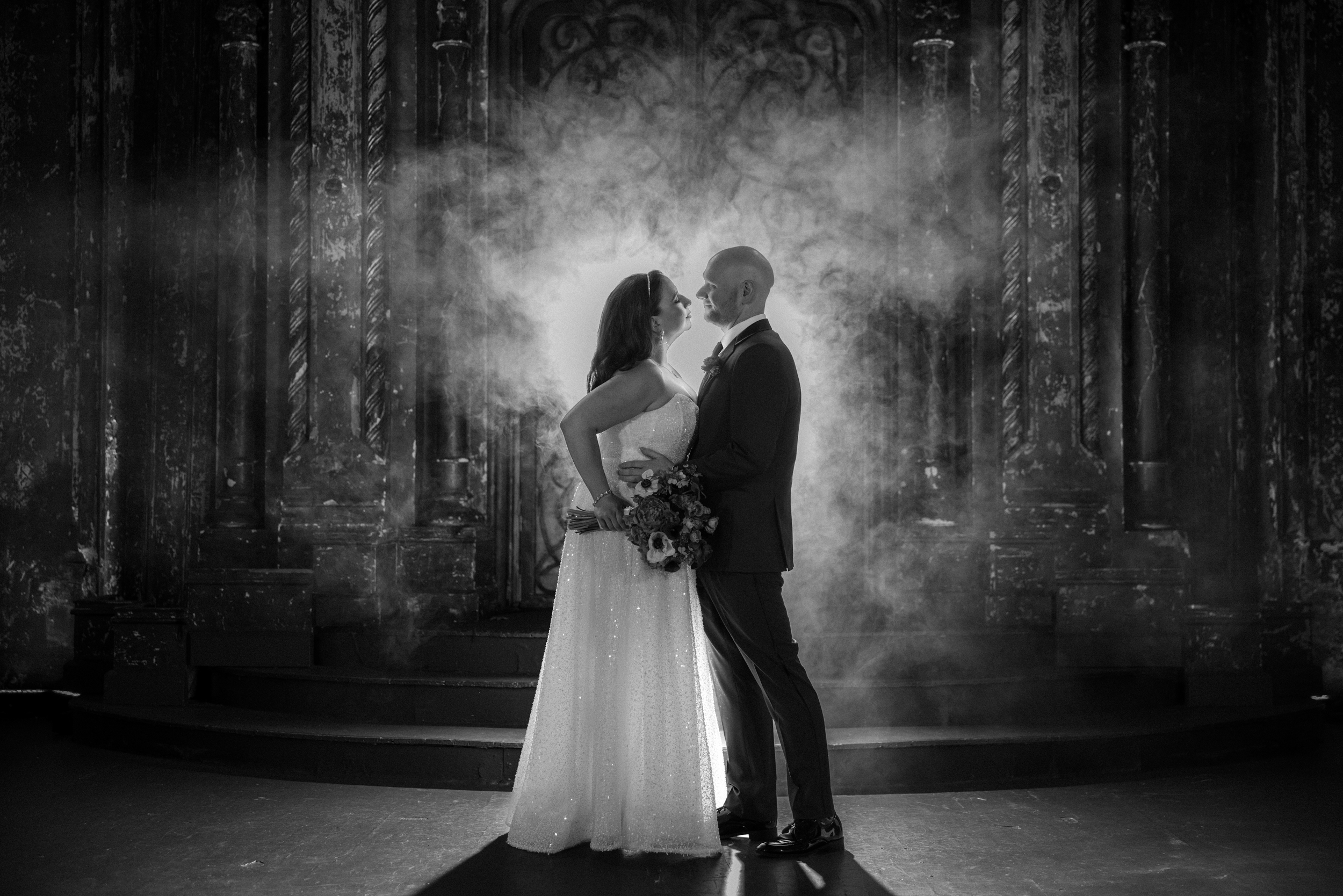 The bride and groom stand facing each other in front of an ornate backdrop at their Angel Orensanz Wedding, surrounded by dramatic lighting and mist, holding a bouquet.