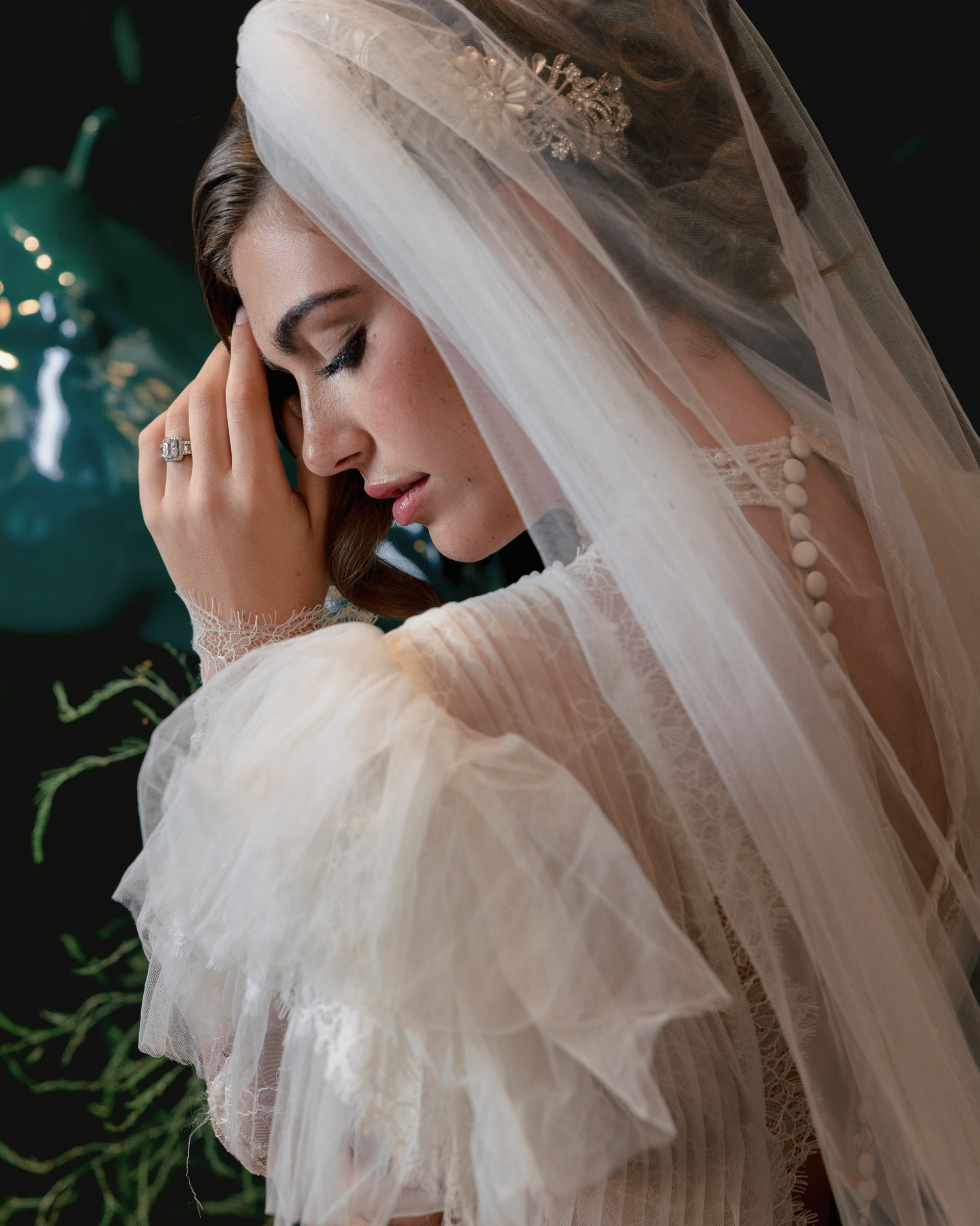 A bride in a white lace dress and veil stands with her hand near her face, eyes closed, showing an engagement ring and a hairpiece—captured in elegant editorial wedding photography in NYC.