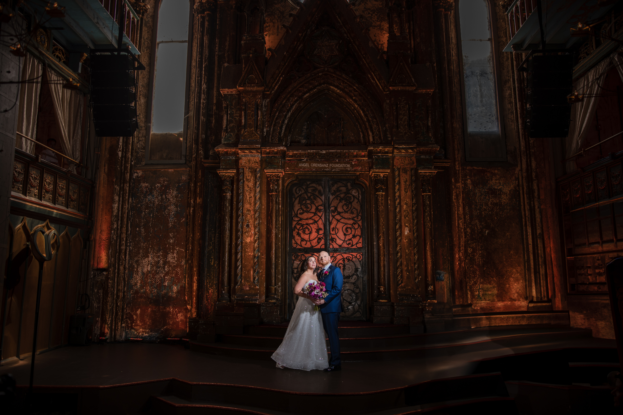A bride and groom stand together holding a bouquet in front of an ornate, dimly lit altar at their unforgettable Angel Orensanz Wedding.