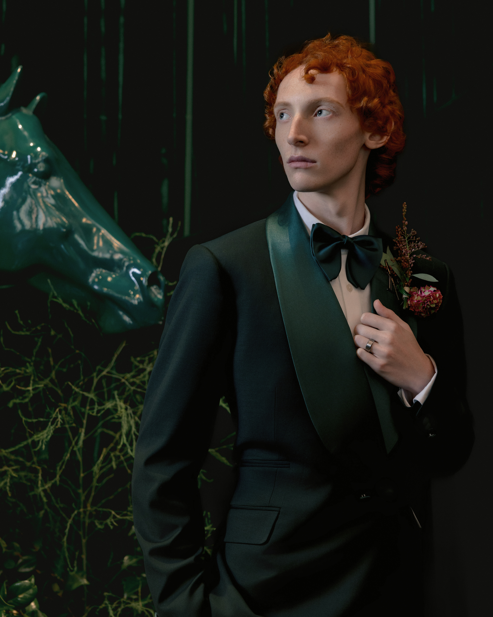 A person with curly red hair in a dark green tuxedo stands in front of a green horse sculpture, holding their lapel with a boutonniere pinned to it—an artistic moment inspired by Editorial Wedding Photography in NYC.