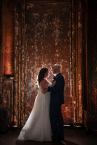 A bride and groom in formal attire stand facing each other, illuminated against a textured, rustic brown backdrop—capturing the timeless elegance of an Angel Orensanz Wedding.