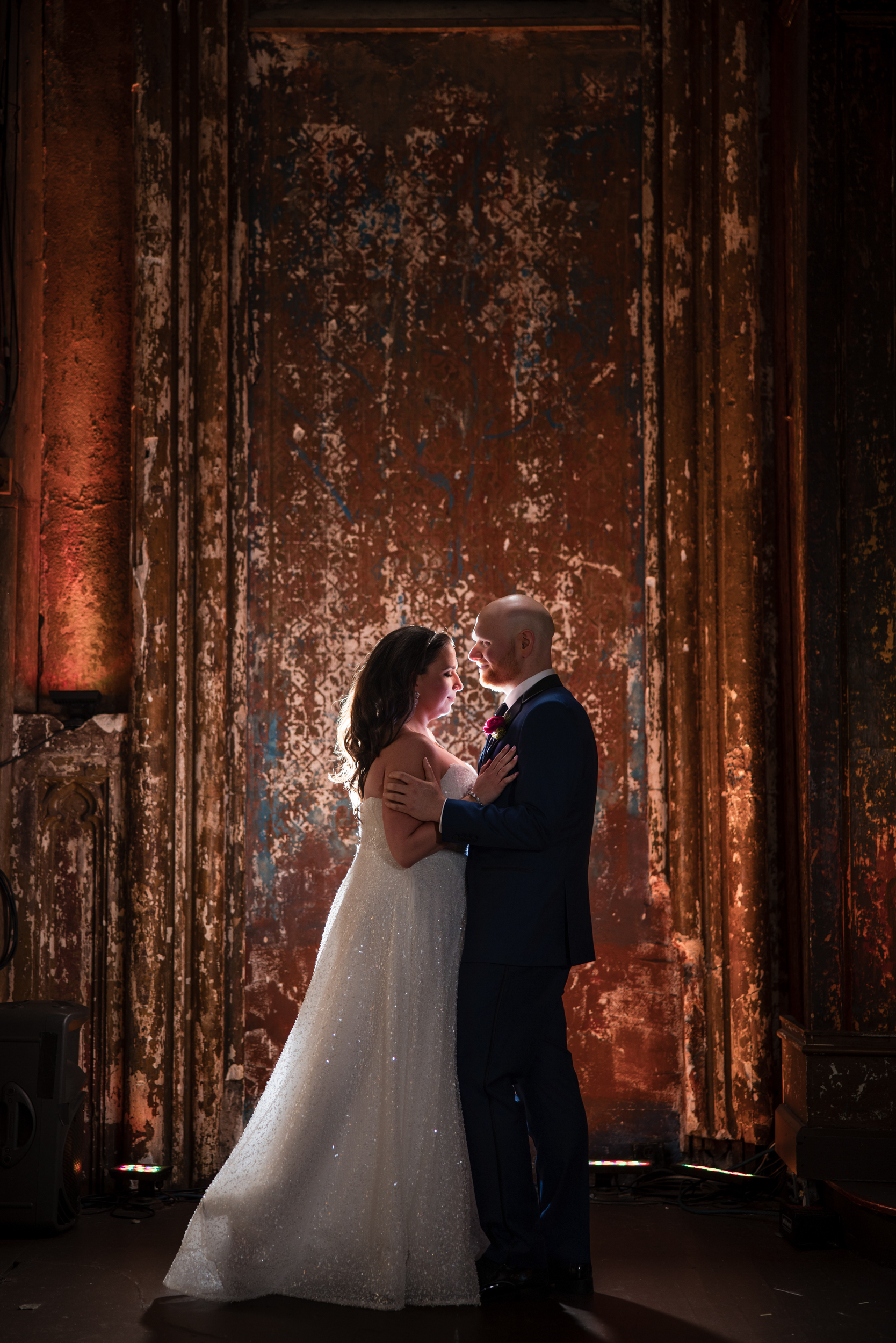 A bride and groom in formal attire stand facing each other, illuminated against a textured, rustic brown backdrop—capturing the timeless elegance of an Angel Orensanz Wedding.