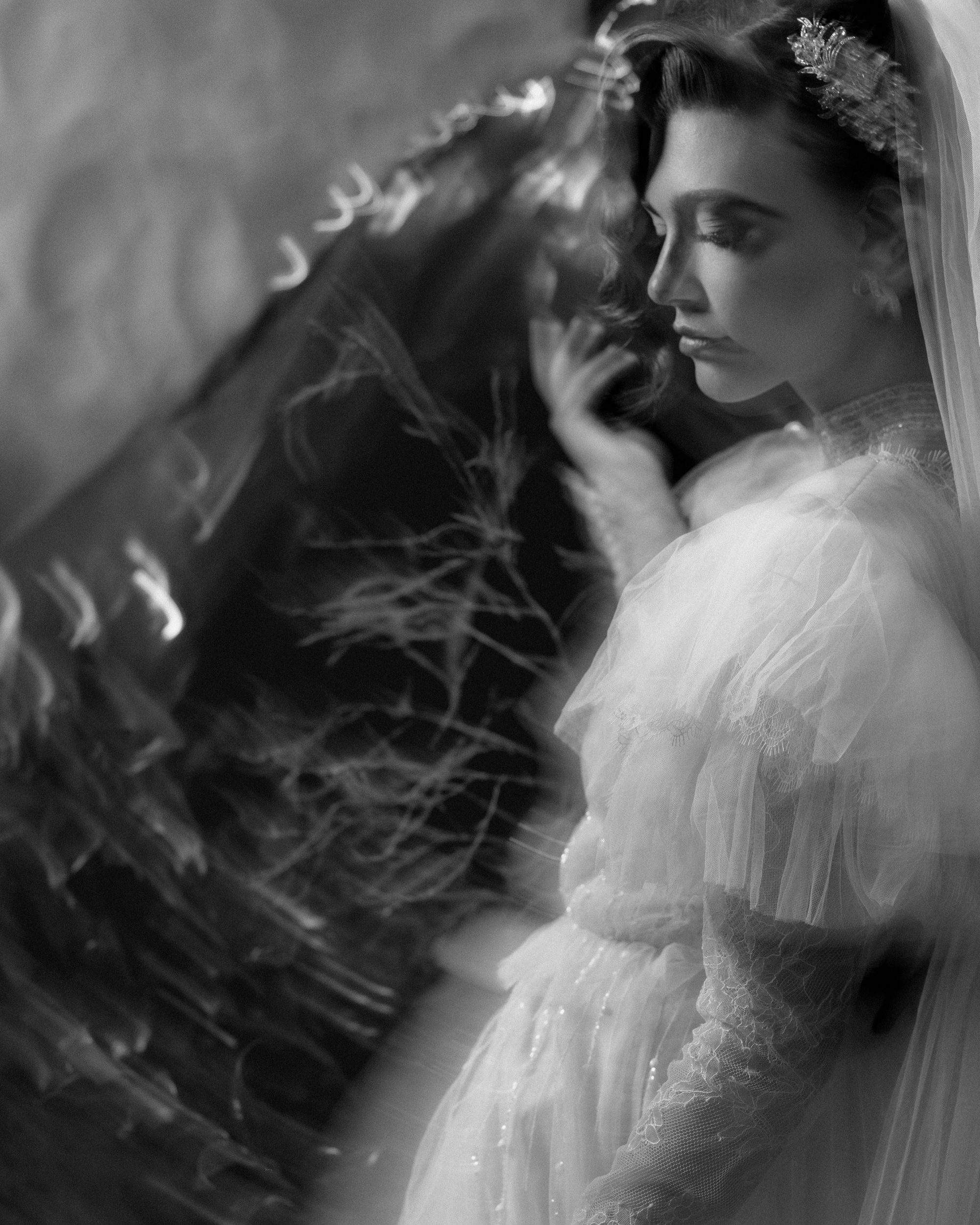 Black and white photo of a woman in a lace dress and veil, standing in profile with her eyes closed and hand raised, surrounded by blurred, abstract light patterns—an elegant example of Editorial Wedding Photography in NYC.