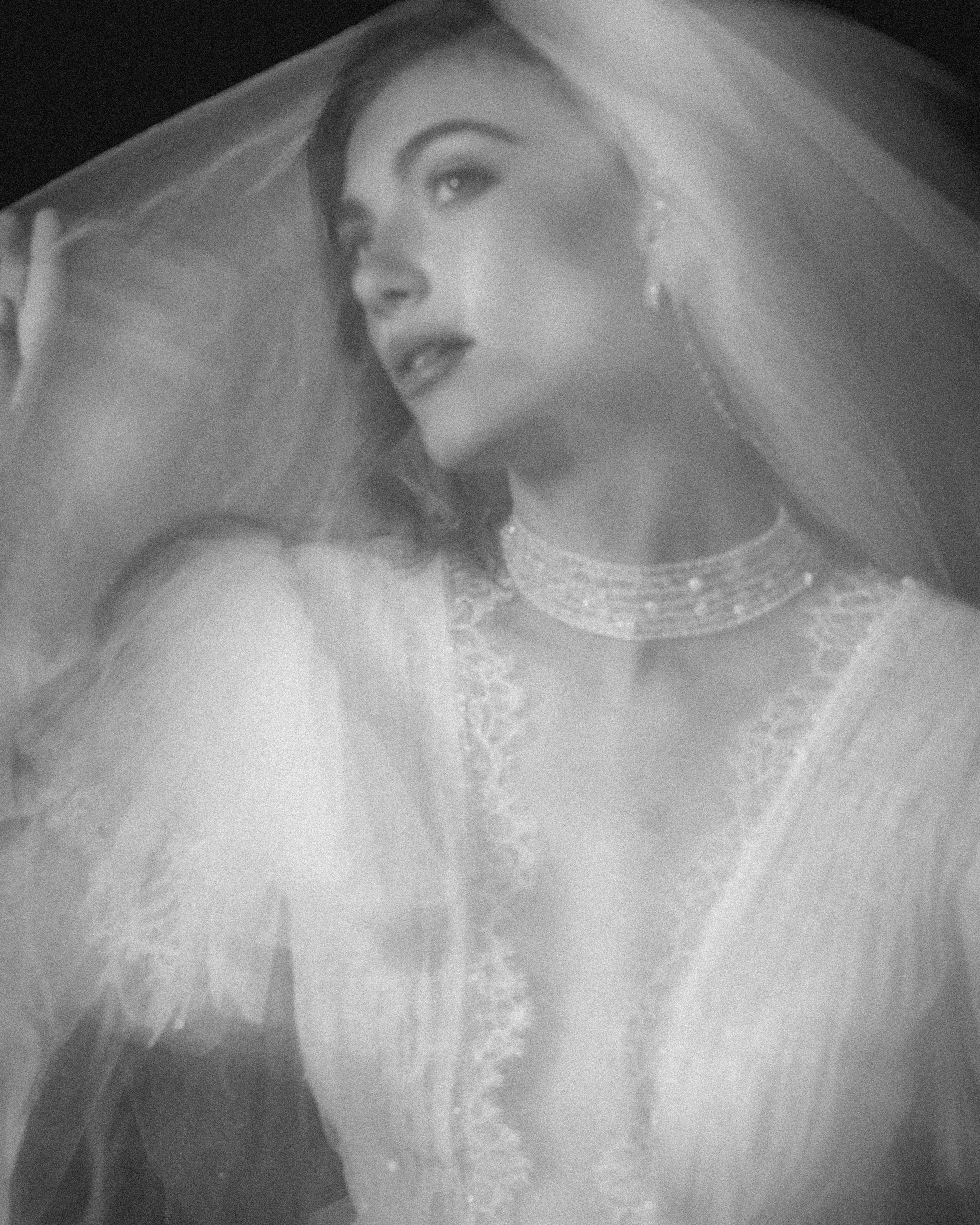 Black and white photo of a woman in a sheer bridal gown and veil, holding part of the veil near her face, with a pearl choker necklace and soft, dreamy lighting—capturing the elegance of editorial wedding photography in NYC.