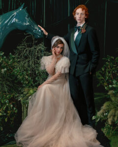 A bride in a tulle gown sits beside a groom in a tuxedo, surrounded by greenery and a teal horse sculpture in a formal, staged setting—perfect inspiration for Editorial Wedding Photography in NYC.