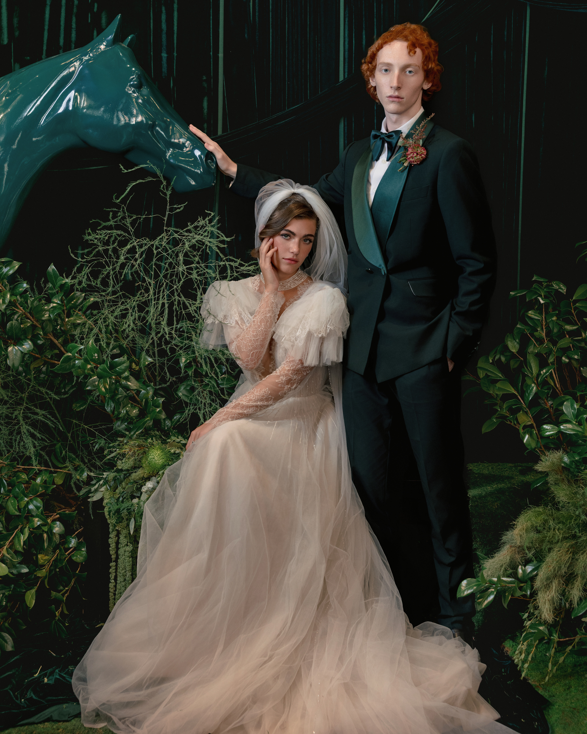 A bride in a tulle gown sits beside a groom in a tuxedo, surrounded by greenery and a teal horse sculpture in a formal, staged setting—perfect inspiration for Editorial Wedding Photography in NYC.