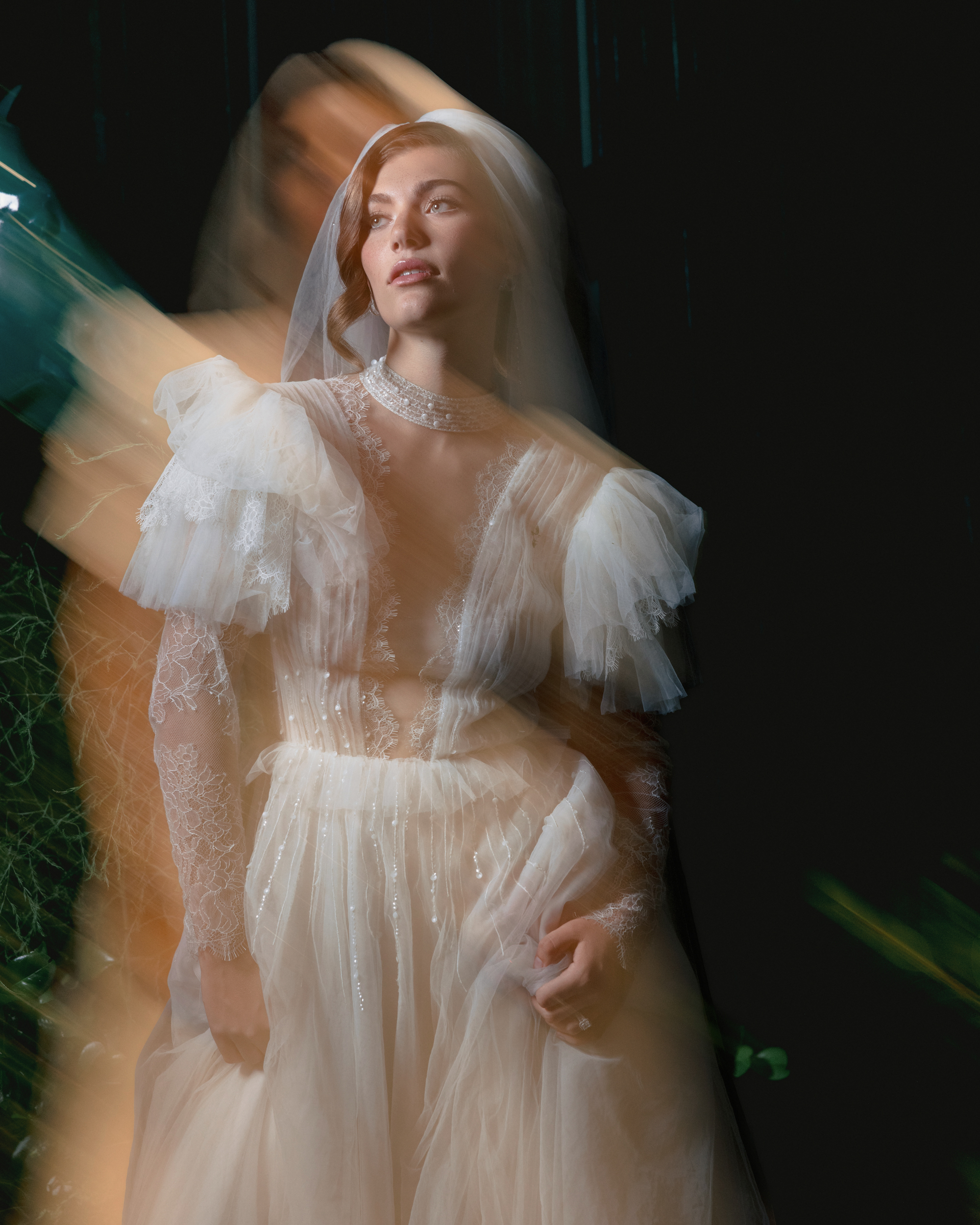A woman in a white bridal gown with lace details stands against a dark background, with motion blur creating a ghostly double image—a striking example of editorial wedding photography in NYC.
