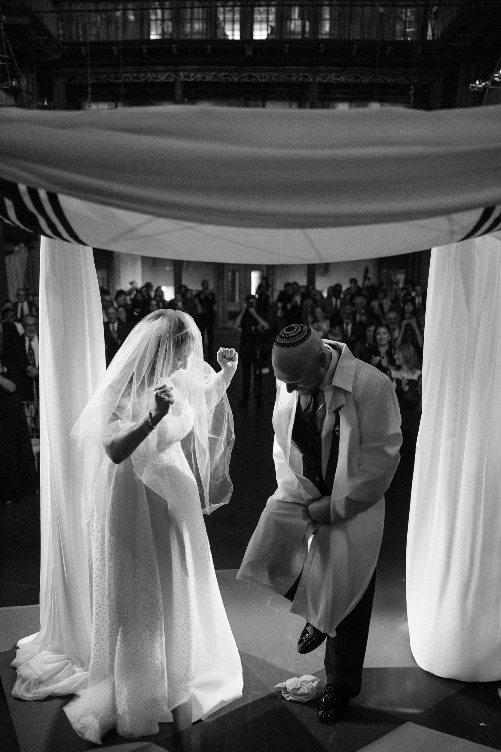 A bride and groom under a wedding canopy at an Angel Orensanz Wedding; the groom is stepping on a glass while the bride looks on, with guests seated in the background.