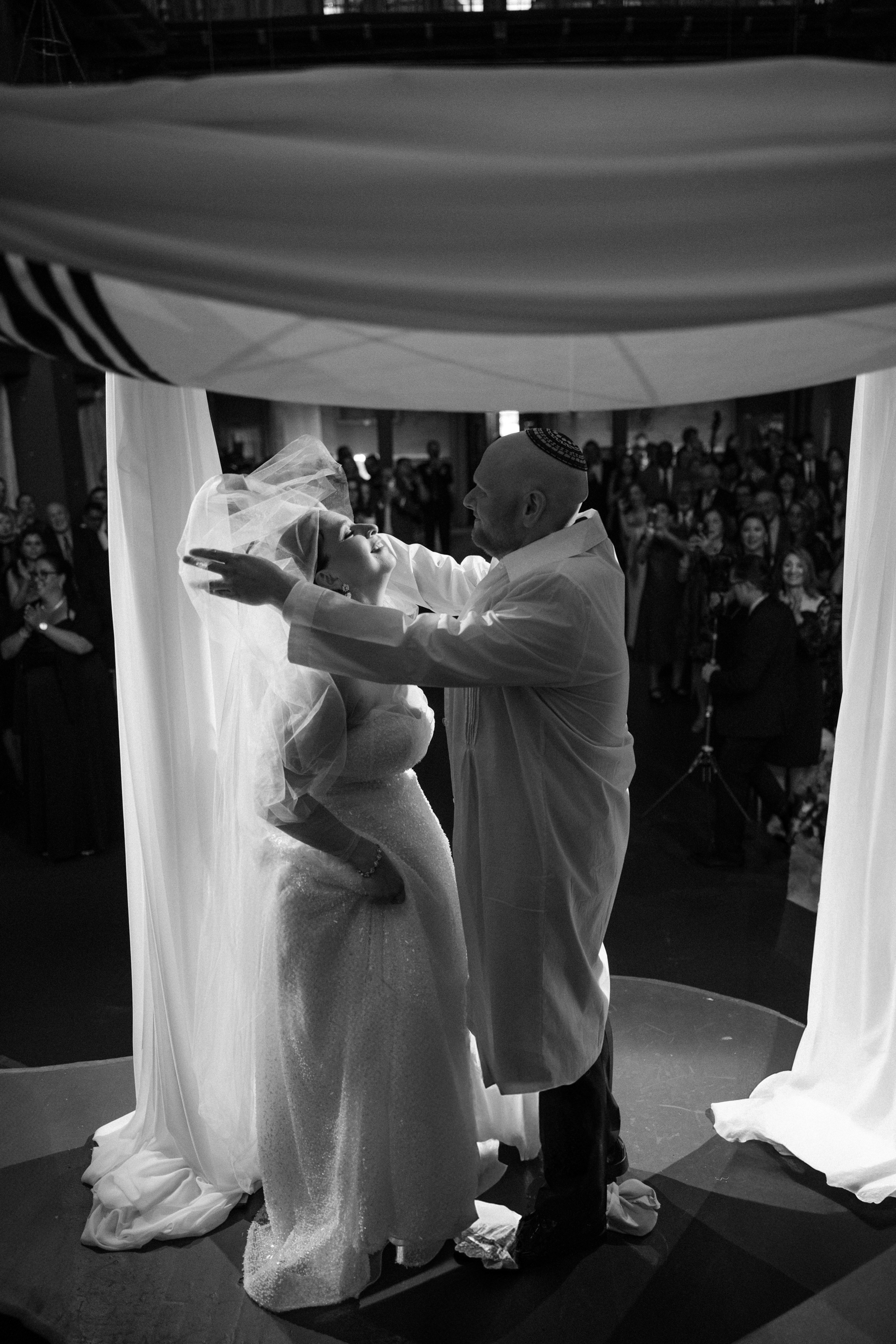 A person lifts a bride's veil under a chuppah during an Angel Orensanz Wedding ceremony, with guests watching in the background.