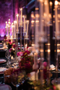 Tall glass candle holders with lit candles and floral centerpieces decorate a formal dining table set with glassware and menus, capturing the romantic ambiance of an Angel Orensanz Wedding in a dimly lit room.