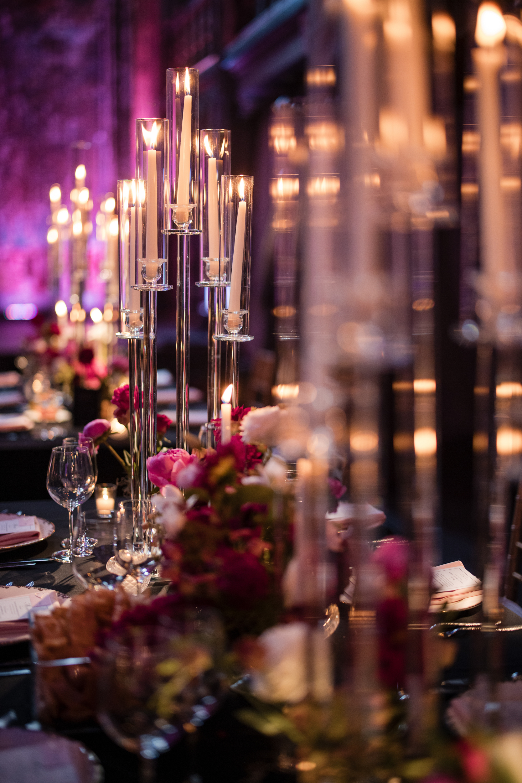 Tall glass candle holders with lit candles and floral centerpieces decorate a formal dining table set with glassware and menus, capturing the romantic ambiance of an Angel Orensanz Wedding in a dimly lit room.