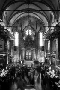 Black and white photo of people gathered in the nave of a grand, gothic-style hall with high vaulted ceilings, arched windows, and ornate details—capturing the timeless elegance of an Angel Orensanz Wedding.