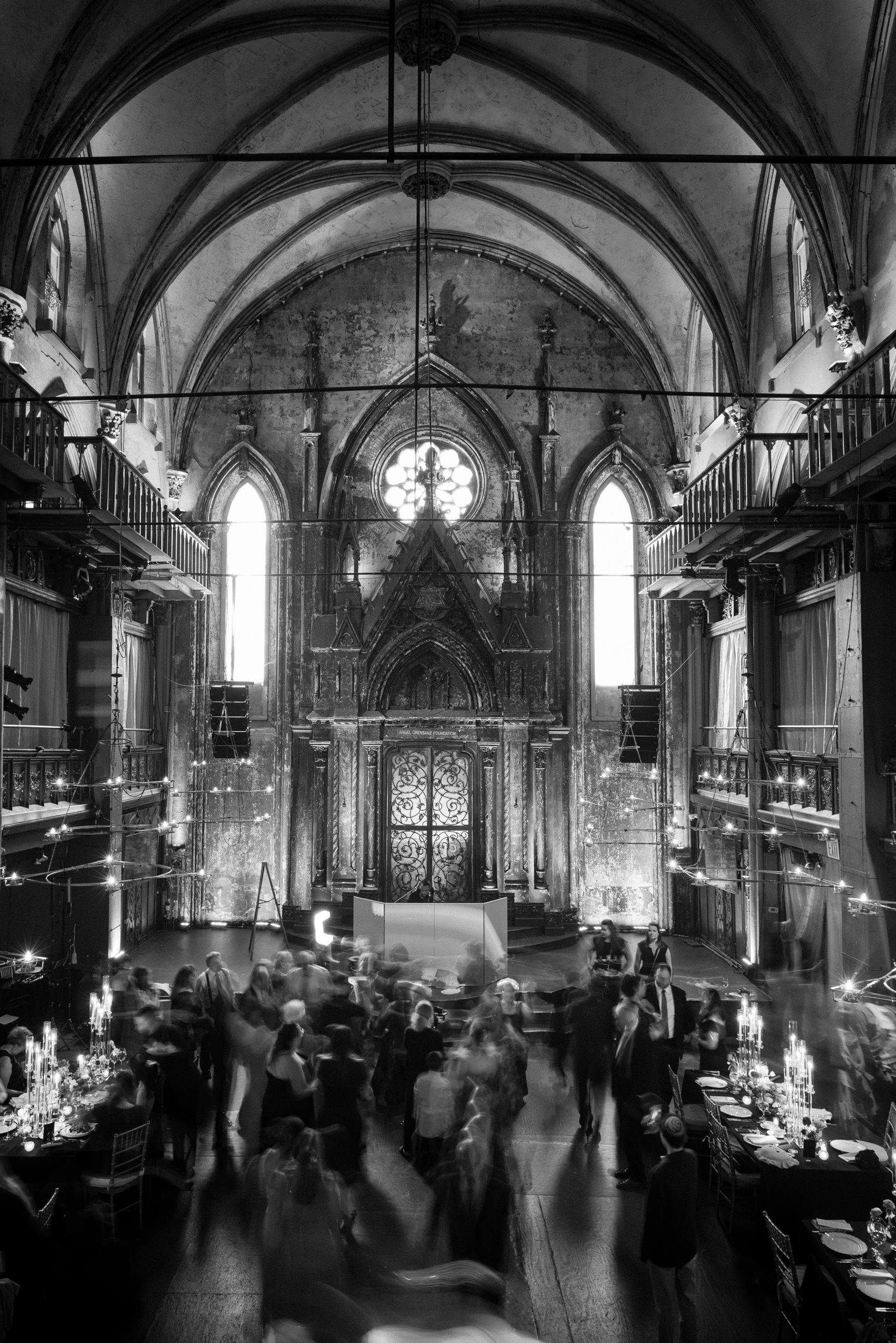Black and white photo of people gathered in the nave of a grand, gothic-style hall with high vaulted ceilings, arched windows, and ornate details—capturing the timeless elegance of an Angel Orensanz Wedding.