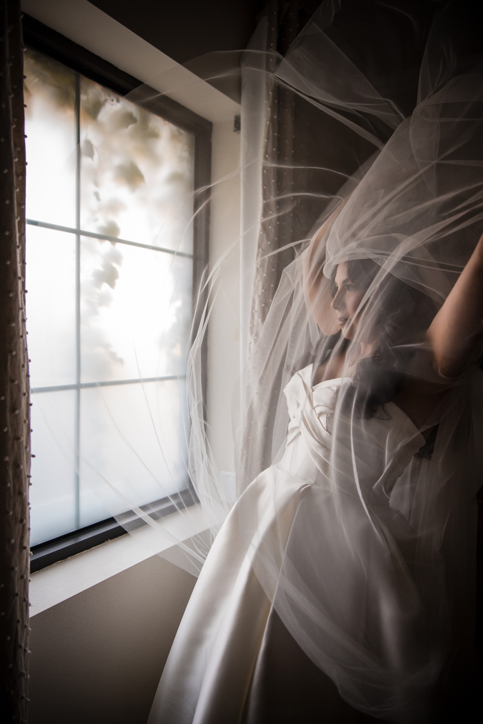 A woman in a white dress stands by a window with sheer fabric billowing around her, reminiscent of a Leoness Cellars wedding, the delicate drapery partially obscuring her figure.