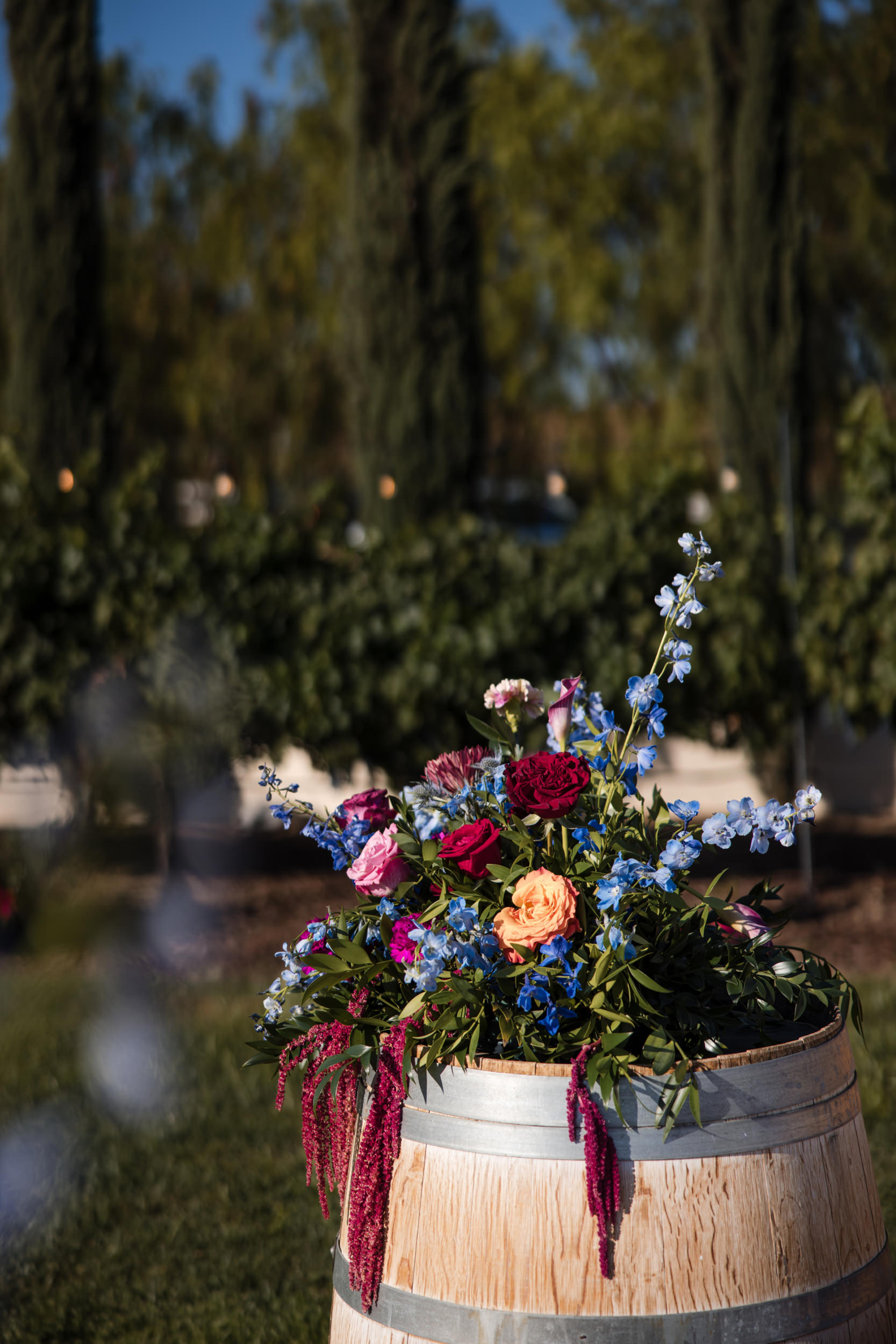 A wooden barrel topped with a colorful floral arrangement sits outdoors near green bushes and tall trees, creating a charming scene reminiscent of a Leoness Cellars wedding.