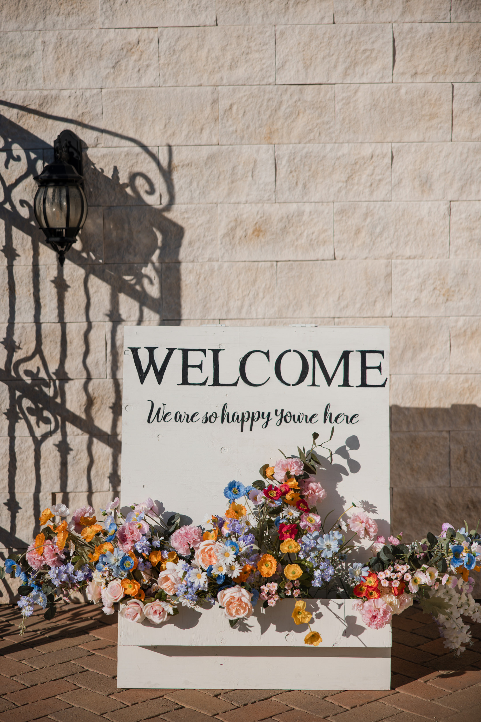 A white sign reading "WELCOME We are so happy you're here" stands against a stone wall at Leoness Cellars wedding, decorated with colorful flowers at the base.