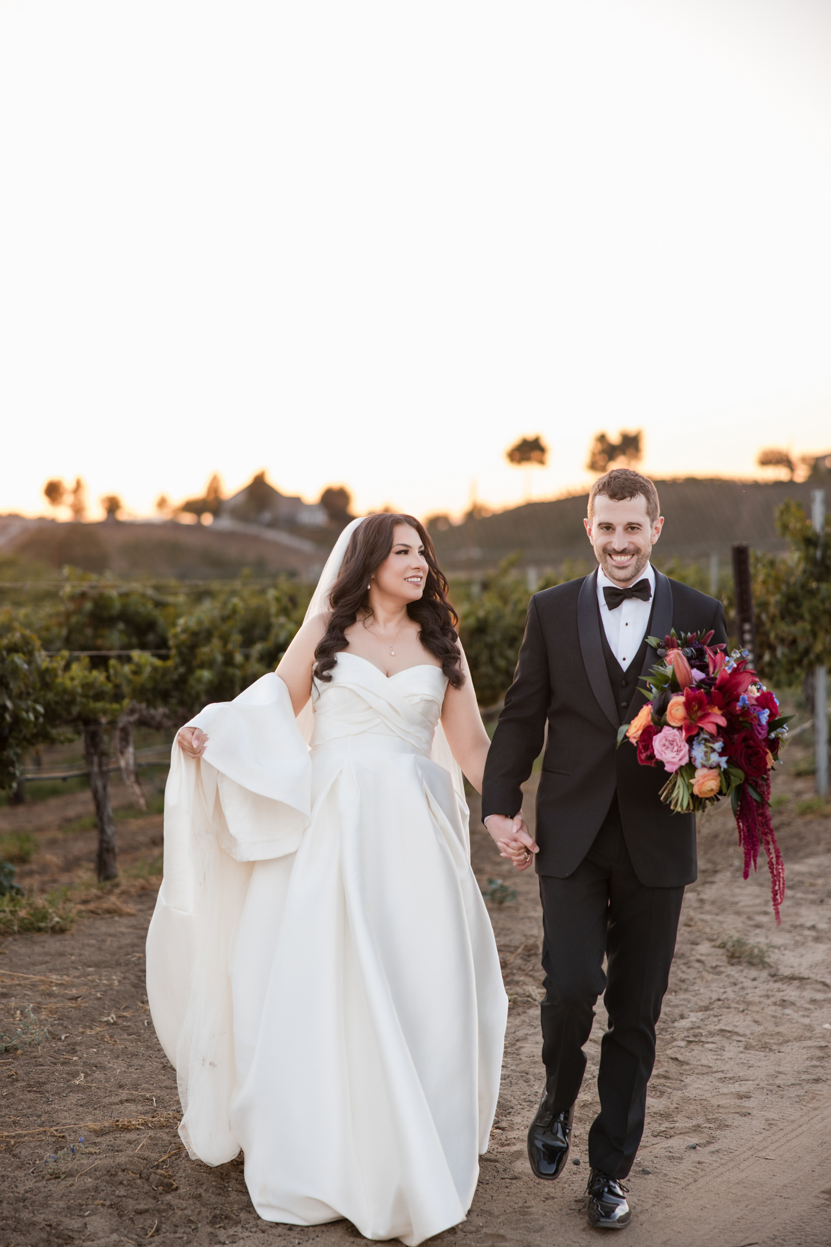 Bride in a white gown and groom in a black tuxedo walk hand-in-hand outdoors at sunset, with the groom holding a colorful bouquet during their romantic Leoness Cellars wedding.