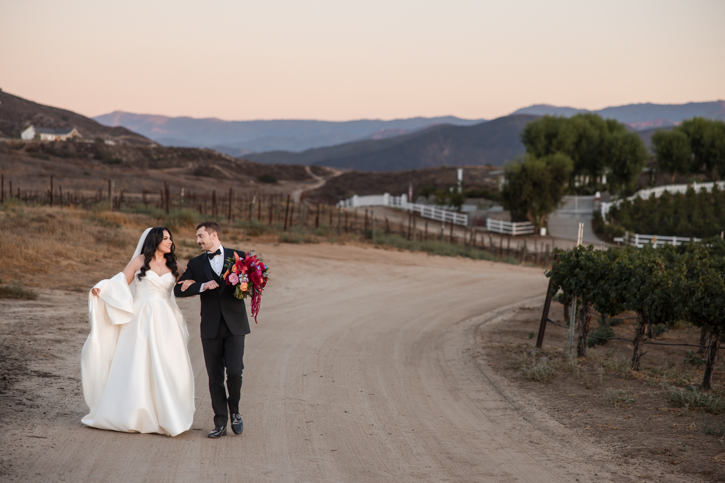 A bride and groom walk together on a dirt road in a rural, vineyard setting at sunset, holding hands and a bouquet of flowers after their romantic Leoness Cellars wedding.