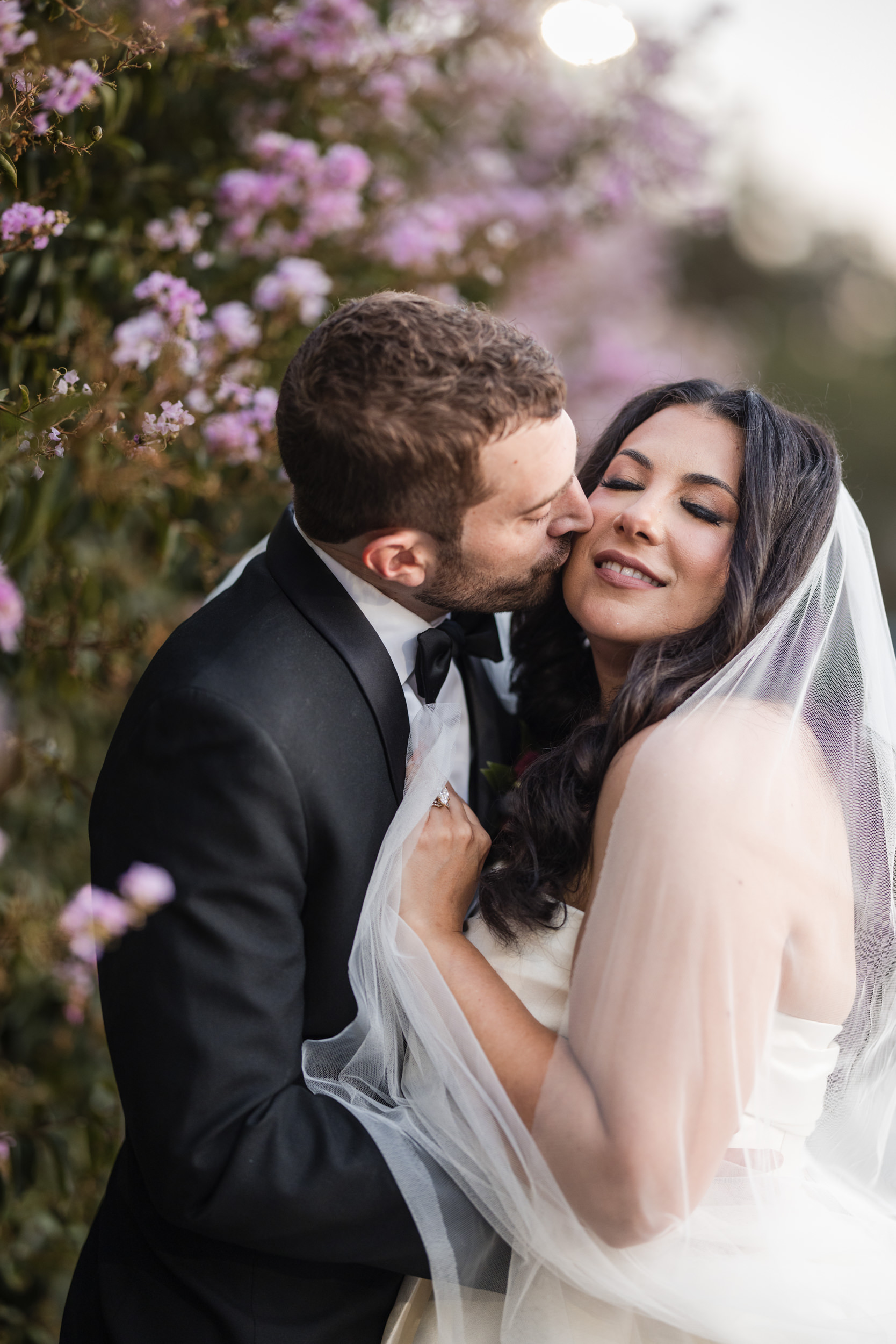 A groom in a black tuxedo kisses a bride in a white dress and veil as they stand close together by blooming purple flowers at their romantic Leoness Cellars wedding.