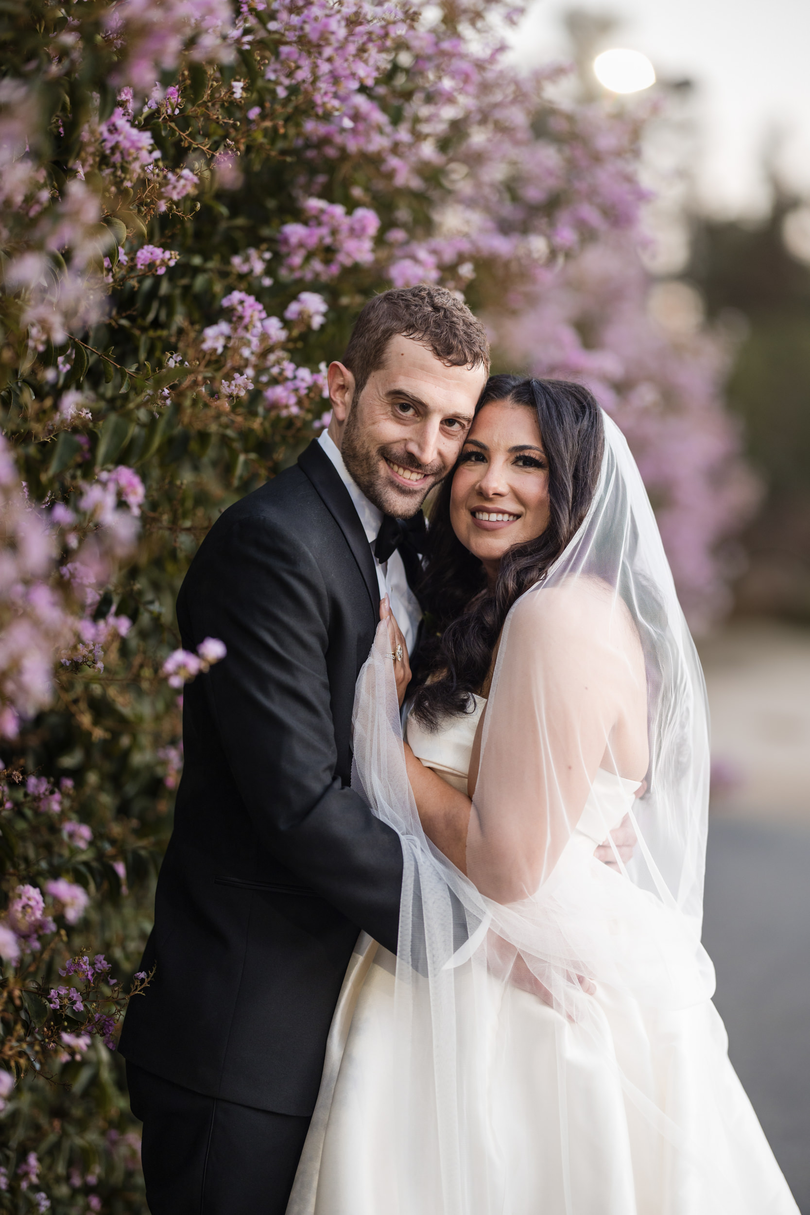 A bride and groom in formal attire stand close together, smiling, with purple flowers in the background at a Leoness Cellars wedding. The bride wears a veil and white dress.