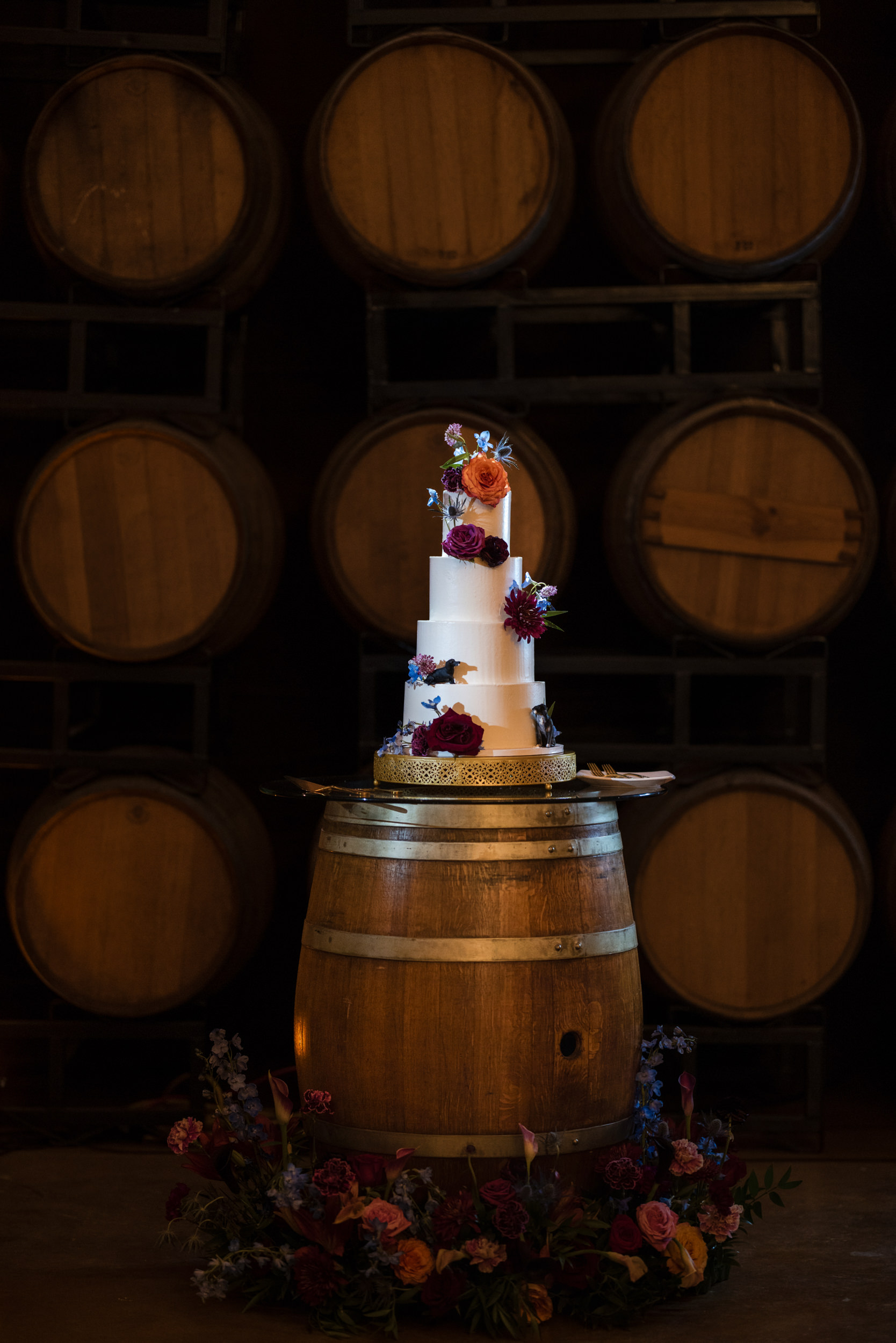 A white, three-tiered wedding cake with floral decorations sits on a wooden barrel at a Leoness Cellars wedding, surrounded by flowers, with stacked wine barrels in the background.