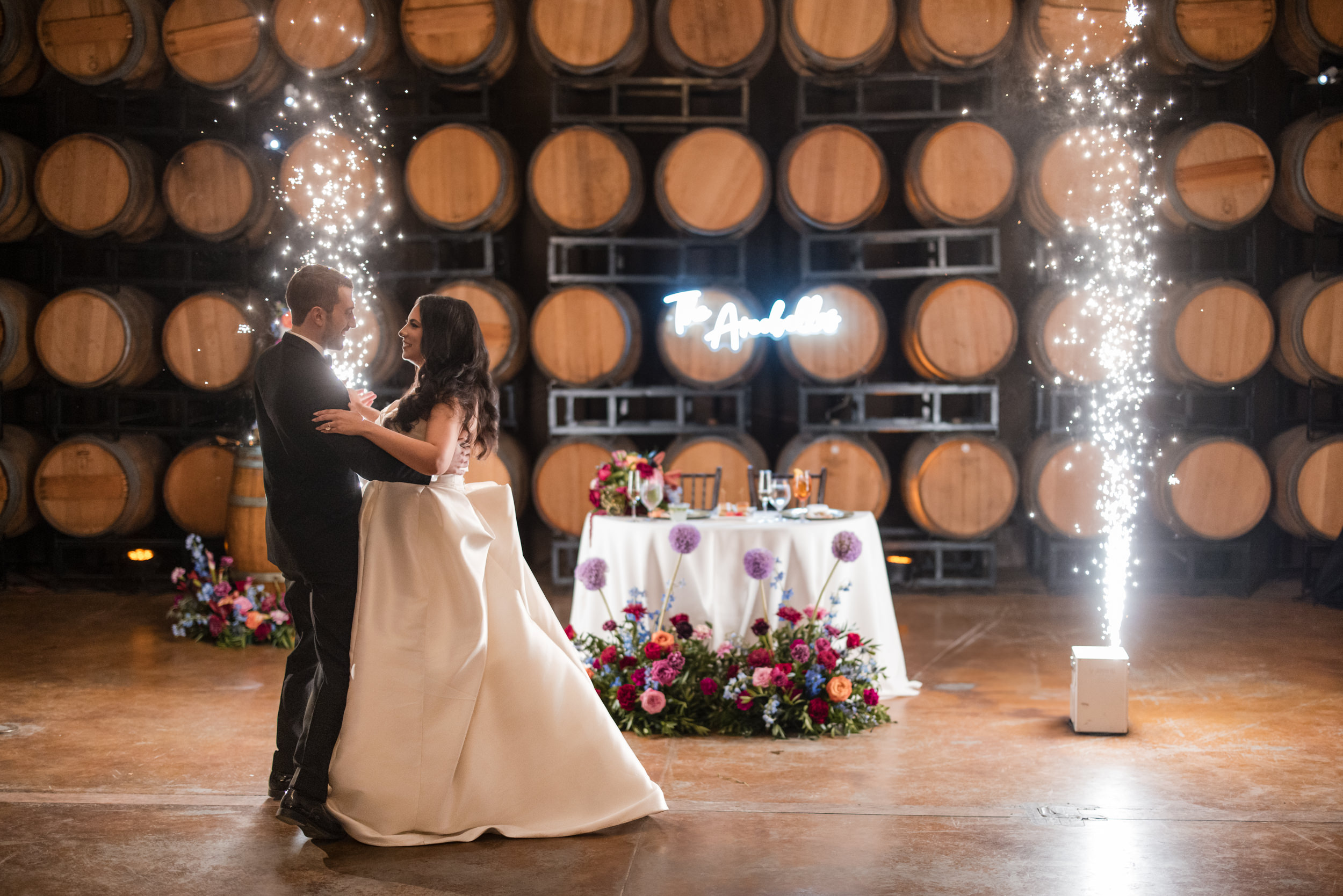 A bride and groom share a first dance in front of a decorated table and wine barrels at their Leoness Cellars wedding, with indoor fireworks sparkling on either side.