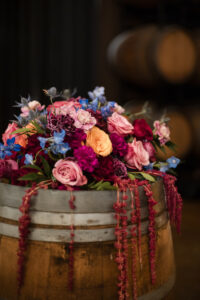 A wooden barrel topped with a colorful arrangement of roses and other flowers in pink, red, yellow, purple, and blue hues creates a romantic Leoness Cellars wedding vibe, with wine barrels blurred in the background.